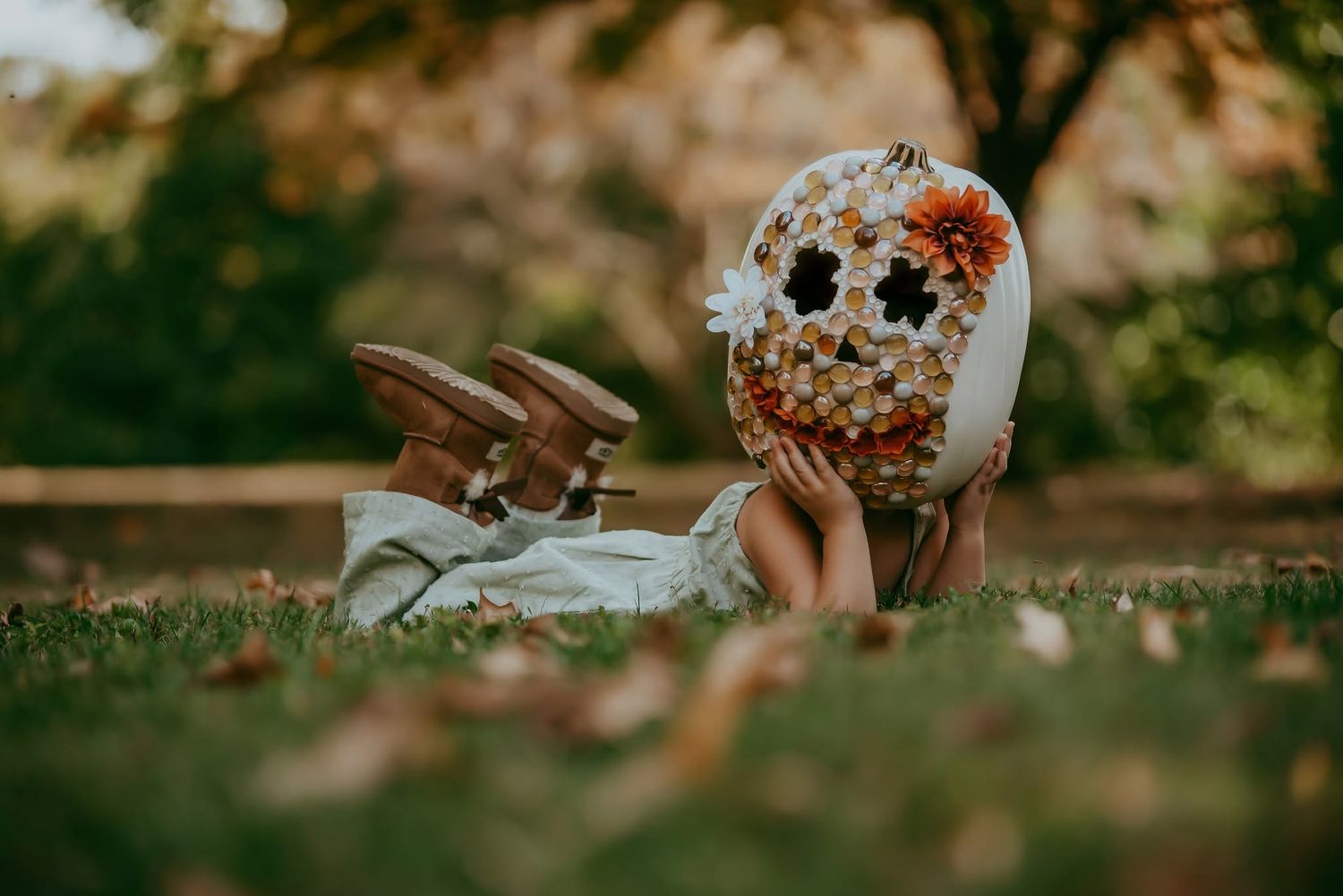 Child lying on grass holding a decorative floral skull pumpkin with brown shoes visible in autumn park setting.