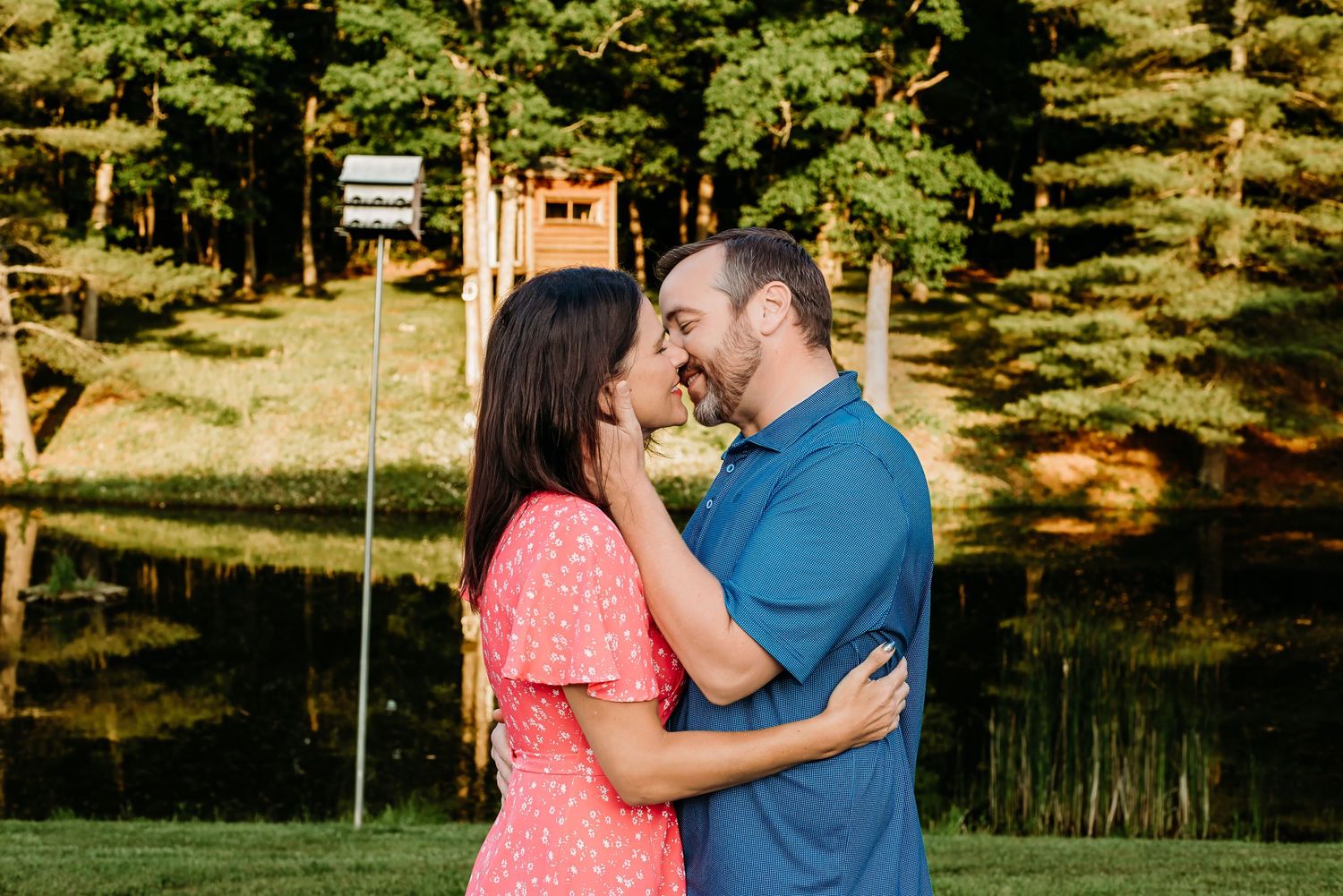 Couple sharing a romantic moment outdoors near a pond surrounded by tall green trees on a sunny day.