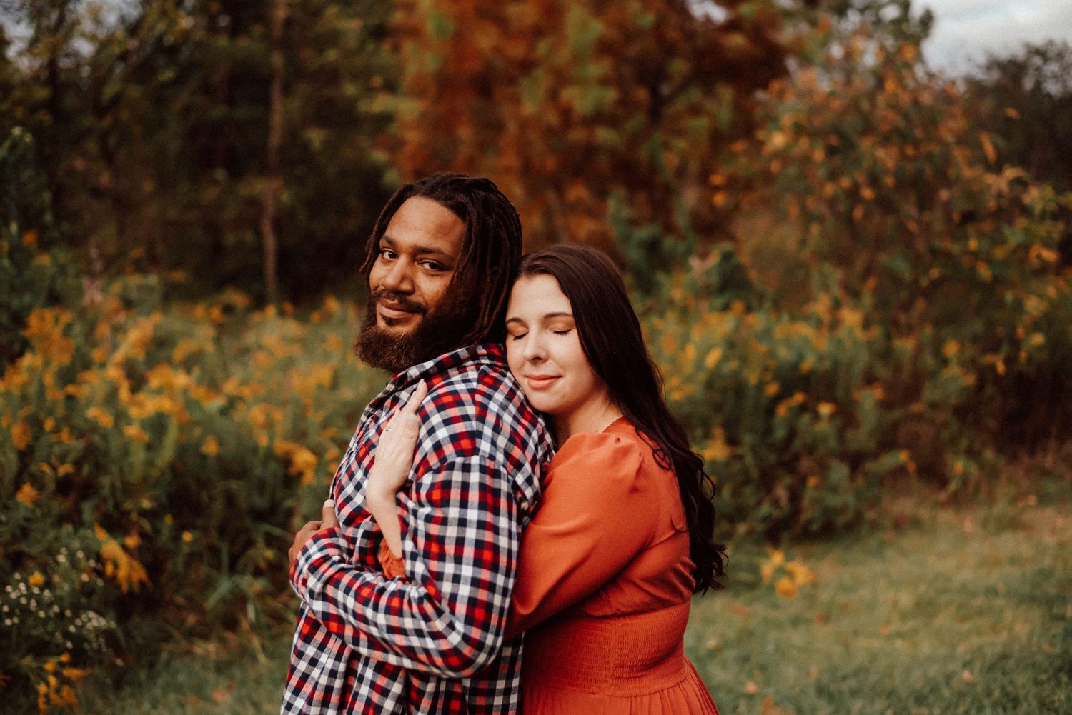 Couple posing outdoors in autumn, man in plaid shirt smiling, woman in orange dress leaning on his back.