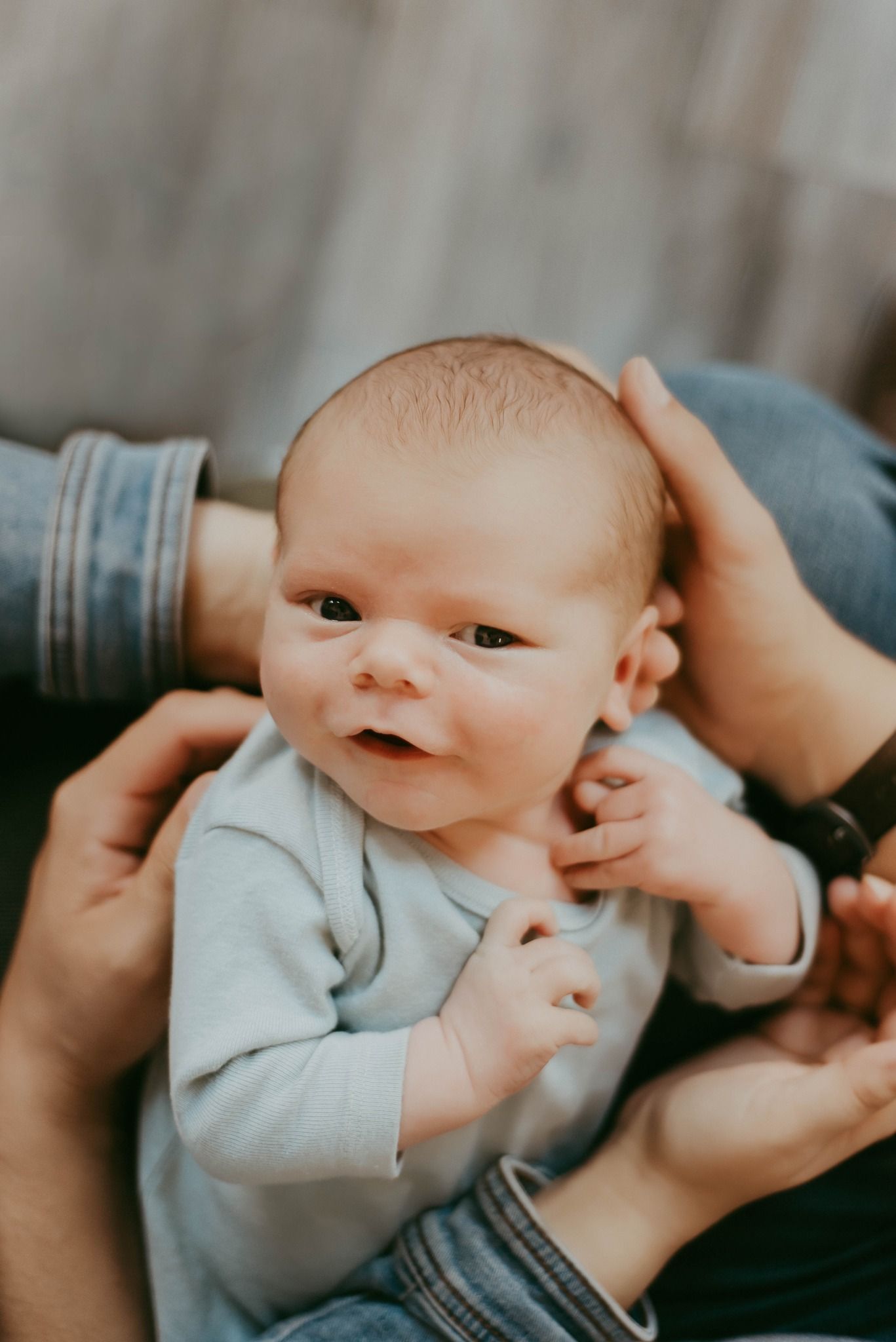 Alert newborn baby in gray onesie looking up at camera with curious expression.