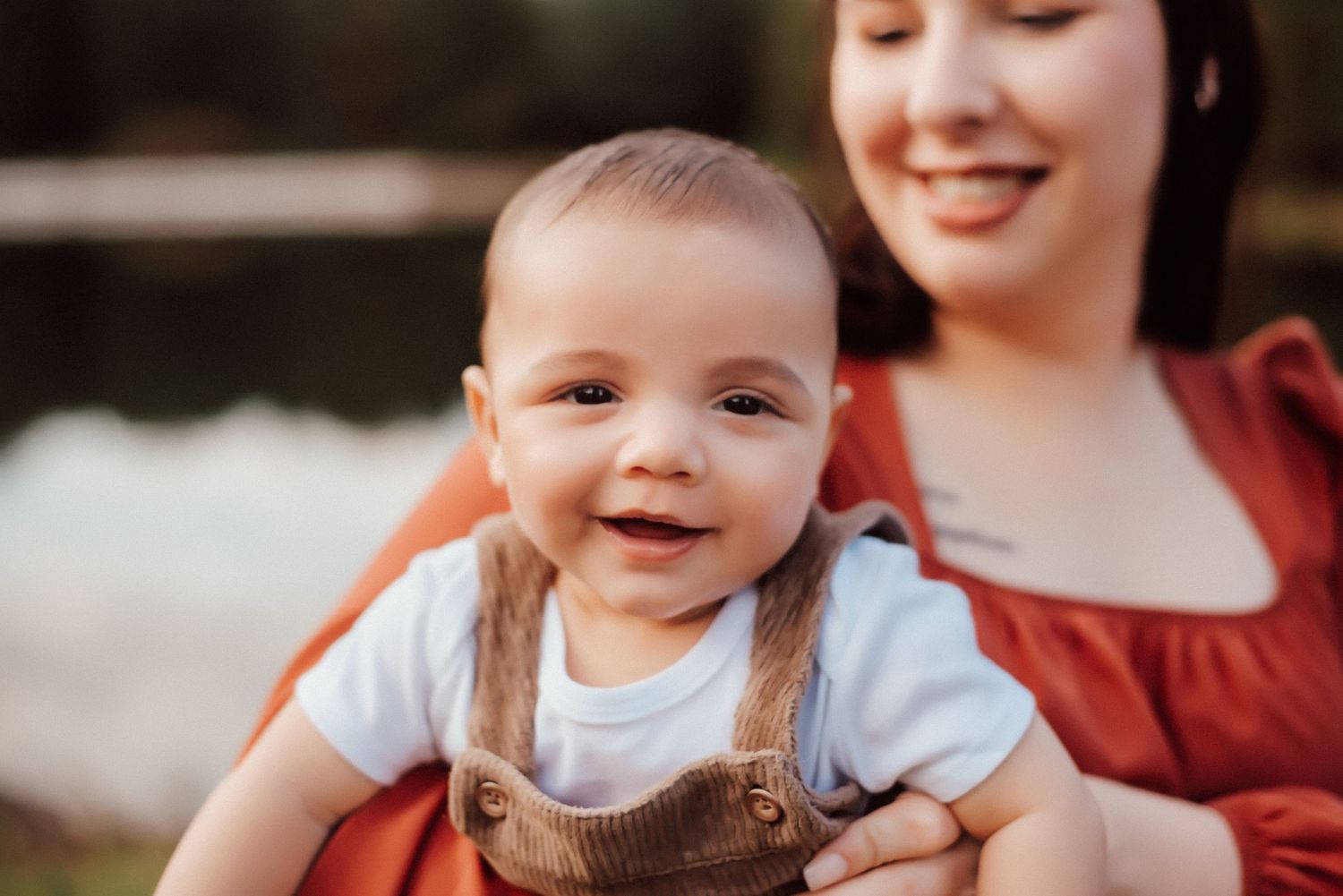 An adorable smiling baby in brown overalls held by a woman in an orange dress outdoors.