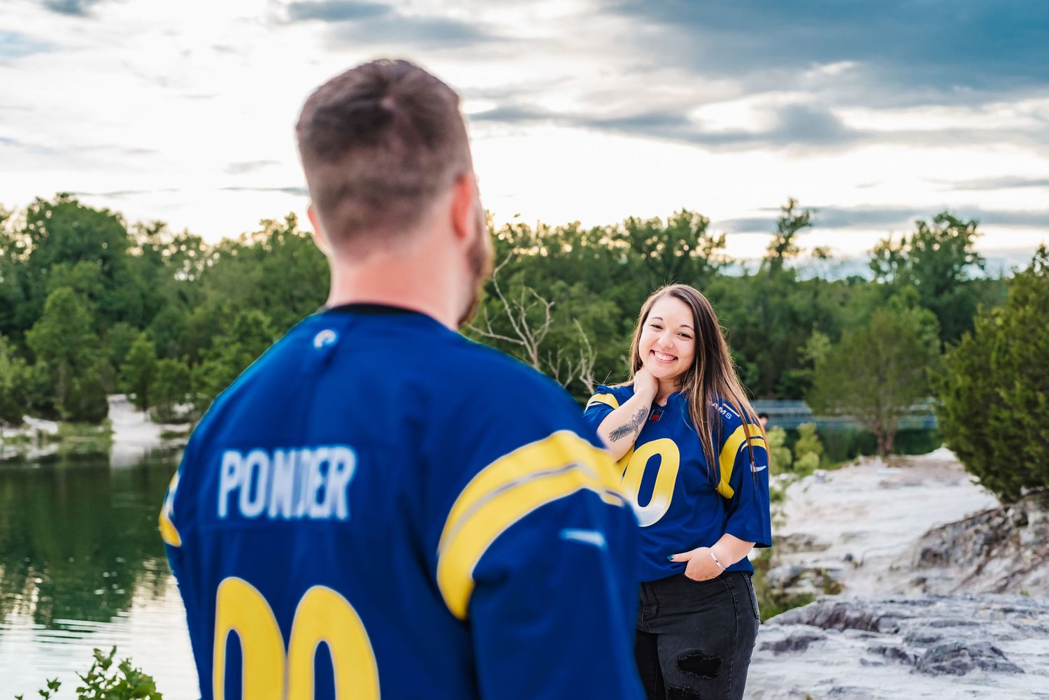 Couple in blue sports jerseys posing by lake at sunset.