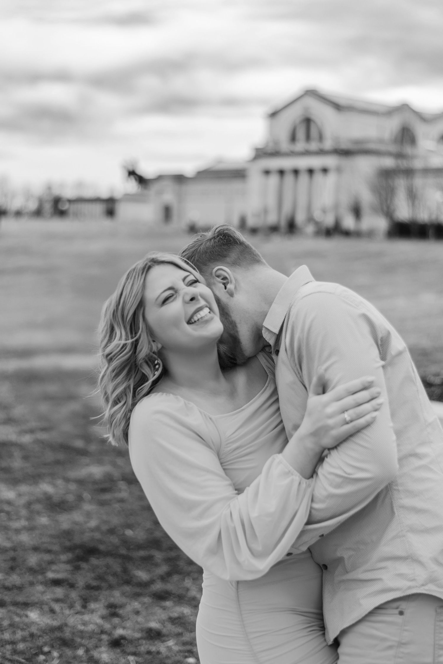 Black and white photo of a smiling woman being kissed on the cheek by a bearded man outdoors near a grand building.