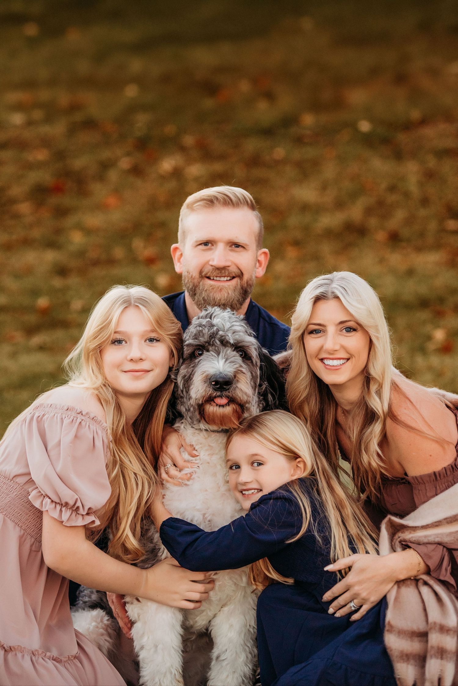 Family of four with a fluffy dog posing outdoors in autumn, wearing coordinated outfits in warm tones.