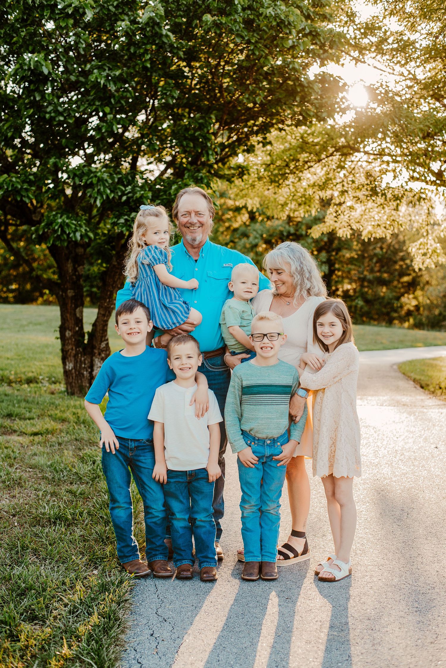 Family of eight posing on a sunlit road surrounded by lush green trees during a warm outdoor photo session.