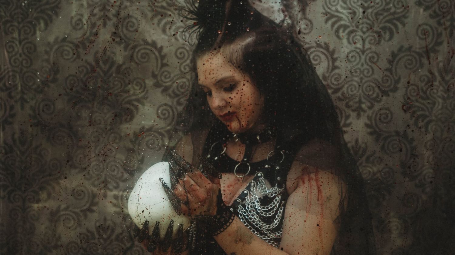 Gothic woman in dark costume holding a white skull with dramatic makeup and vintage wallpaper backdrop.