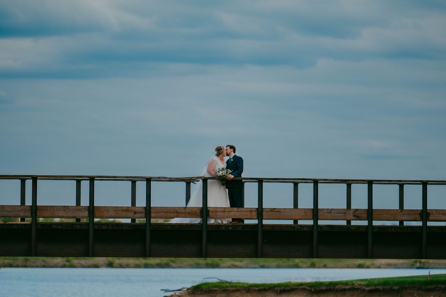 A couple shares an intimate moment on a wooden bridge overlooking water under blue skies.