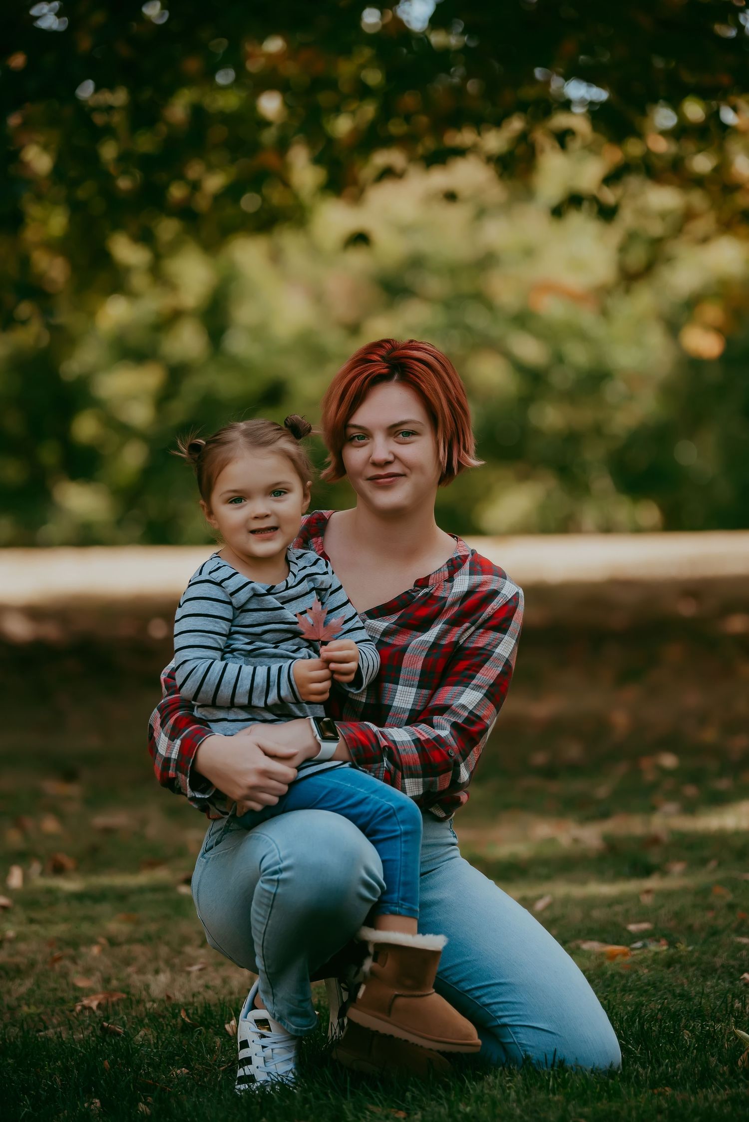 A young woman with red hair holds a toddler girl on her lap while sitting on grass in a park.
