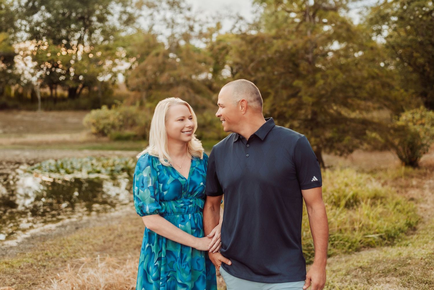 Couple shares loving moment by pond during sunset with woman in teal dress and man in navy shirt.