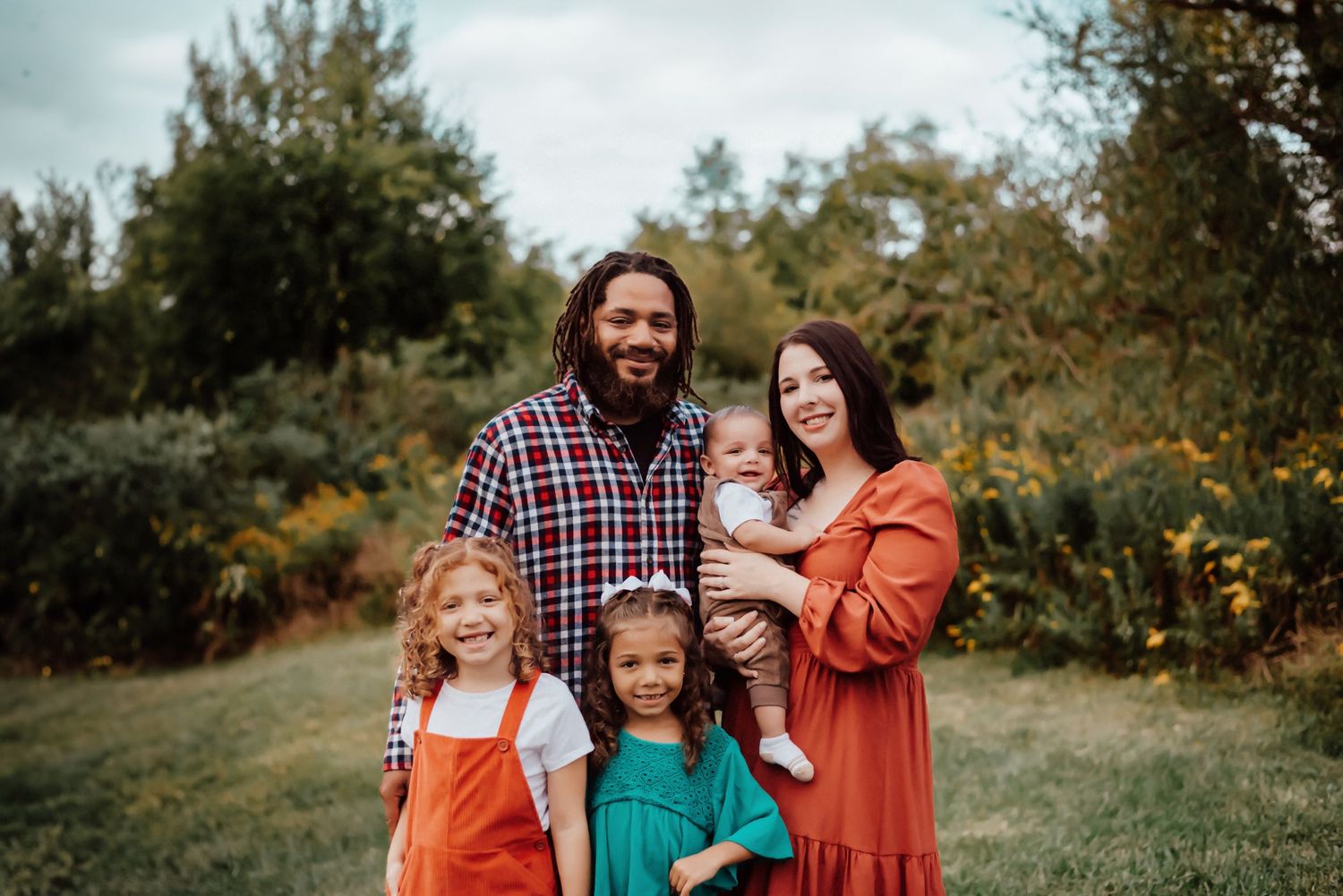 A happy family of five poses outdoors in fall, parents holding a baby with two young girls in colorful dresses.