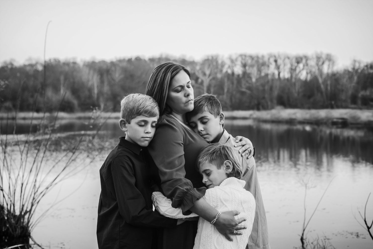 Black and white portrait of family embracing by lakeside with trees in background.
