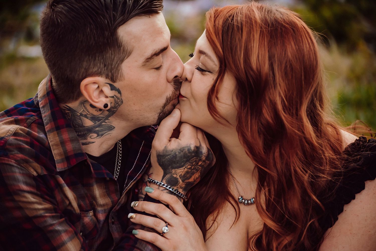 Tattooed couple sharing a romantic kiss outdoors, man with neck tattoos and woman with long red hair.