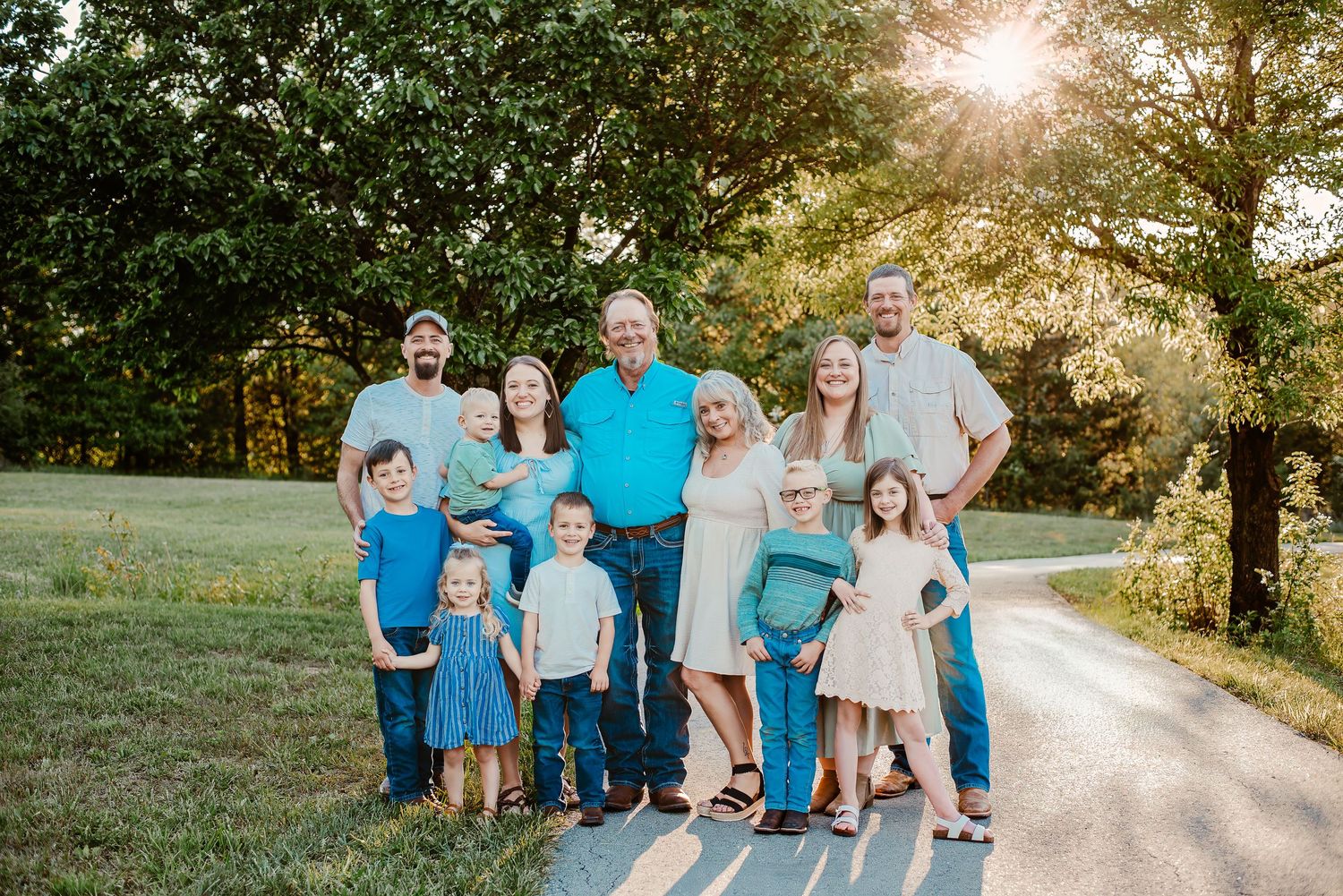 Large family portrait outdoors at golden hour, adults and children in coordinating blue and white outfits on a tree-lined path.