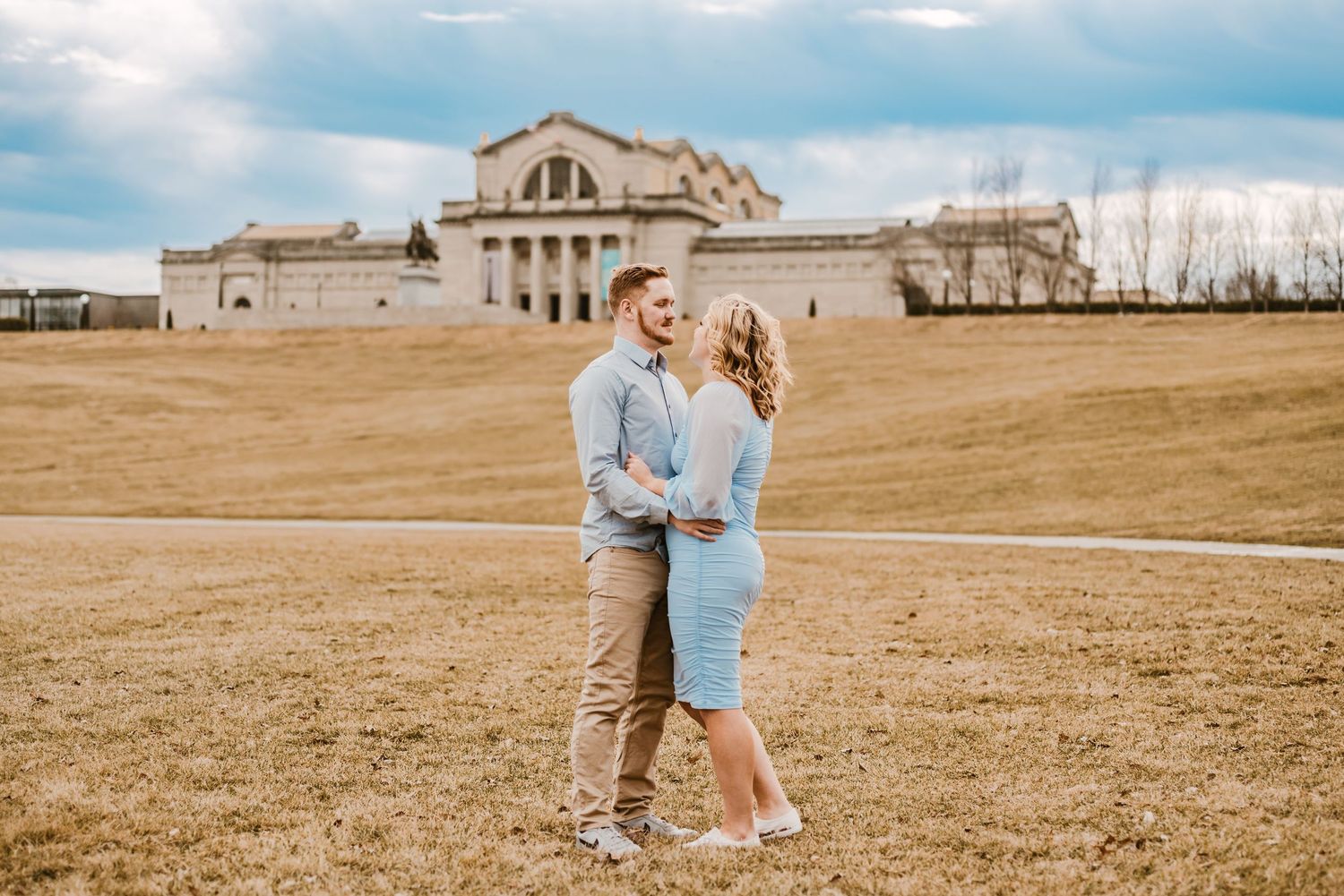 A couple shares a romantic moment on a grassy hill with a grand historic building in the background.