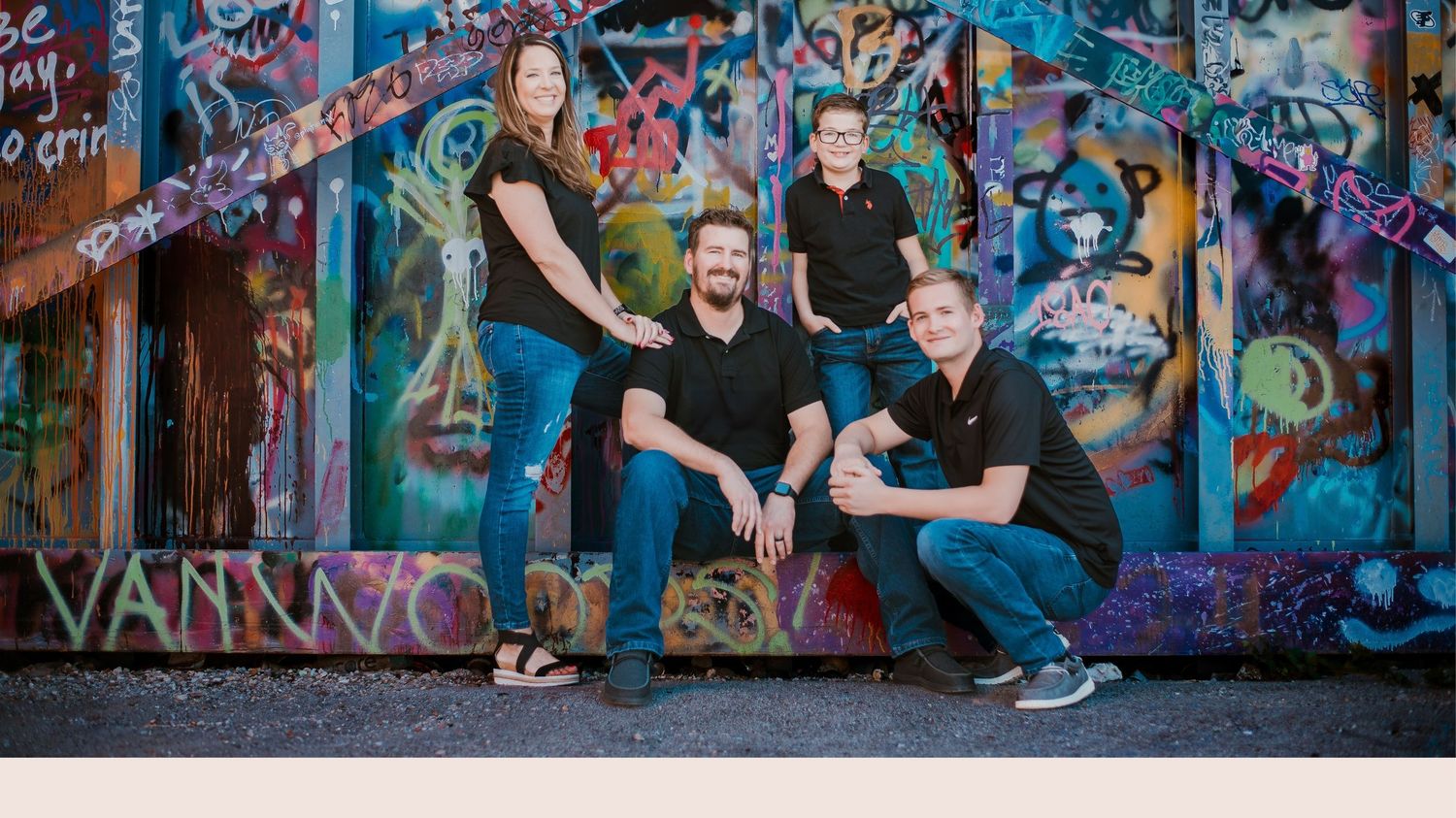 Four people in black shirts pose in front of a vibrant graffiti wall outdoors.