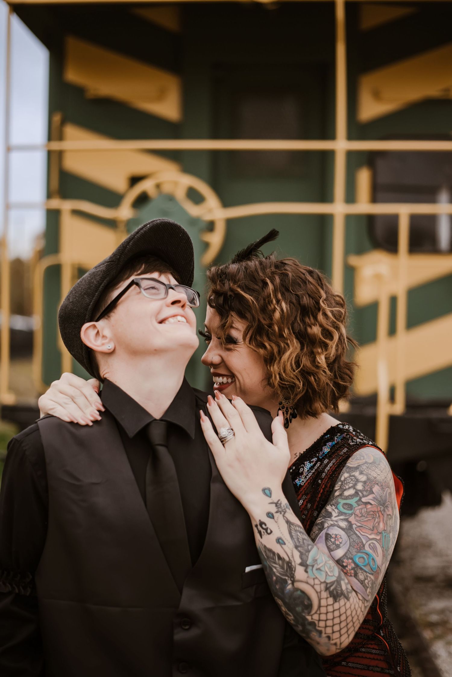 Two people laughing and embracing outdoors, one with tattoos and curly hair, the other in a black suit and cap, near a train.
