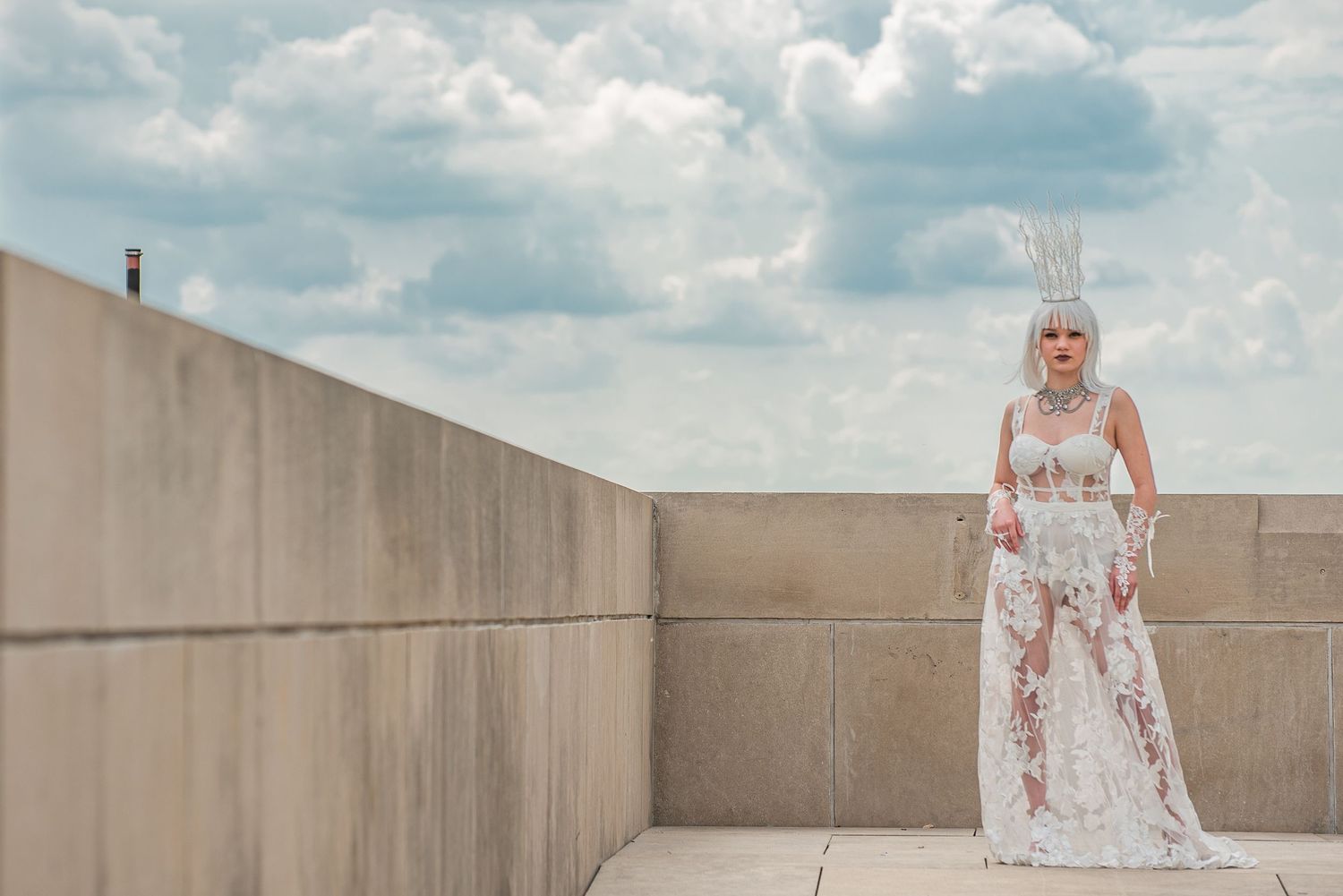 Bride in white lace gown poses dramatically on rooftop under cloudy sky.
