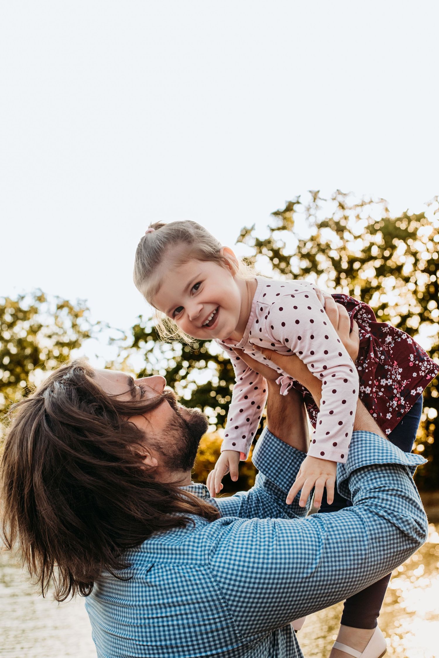 A person in a blue checkered shirt lifts a small child wearing polka dots during a sunset photo session outdoors.