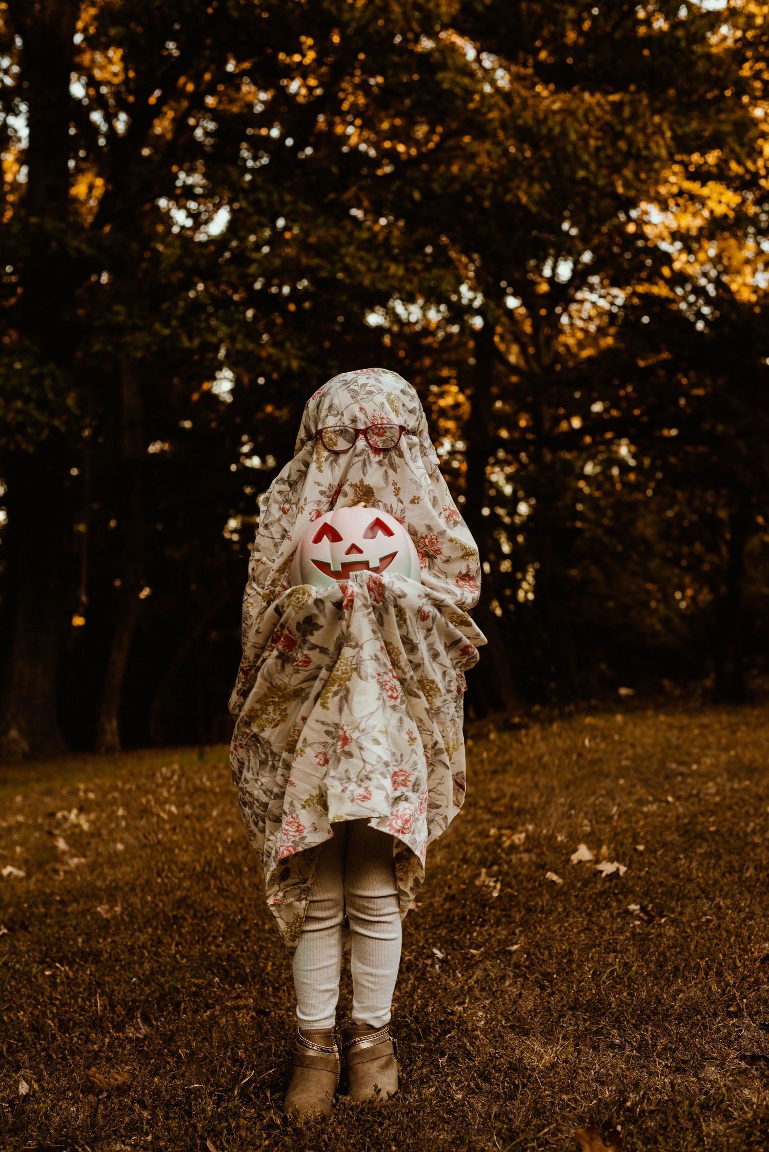 Child wearing ghost costume with jack-o-lantern face standing in autumn forest.