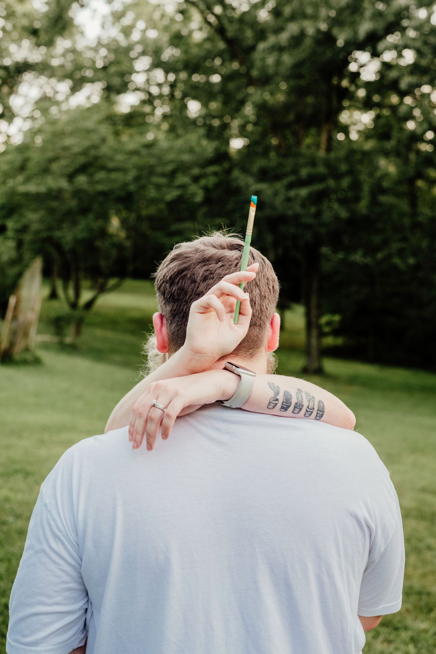 A couple embraces outdoors in a lush green garden setting wearing light colored clothing.
