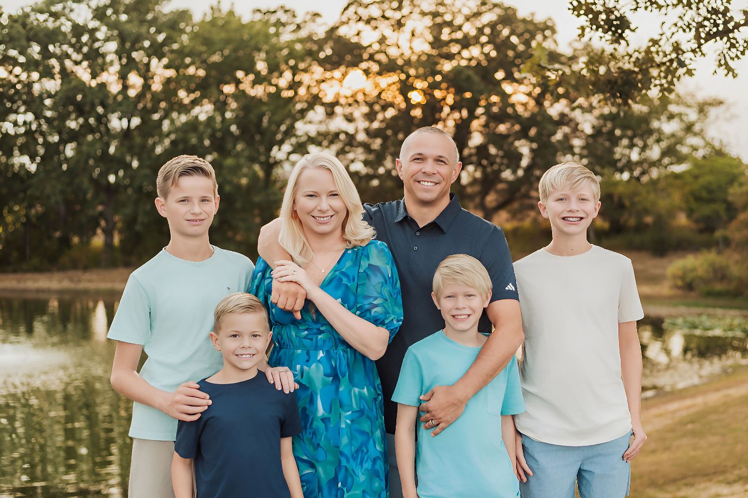 A smiling family of six poses outdoors near a pond at golden hour, wearing coordinated blue and teal outfits.