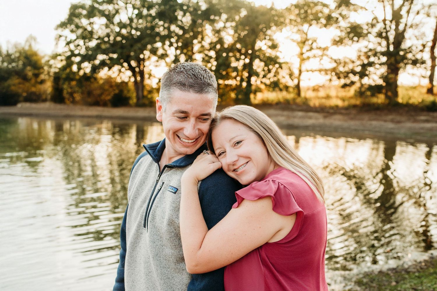 Happy couple embracing and smiling by a sunlit lake surrounded by autumn trees.