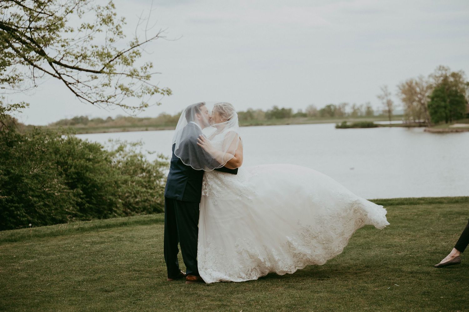 Wedding couple embracing outdoors by a lake with flowing white dress in the wind.