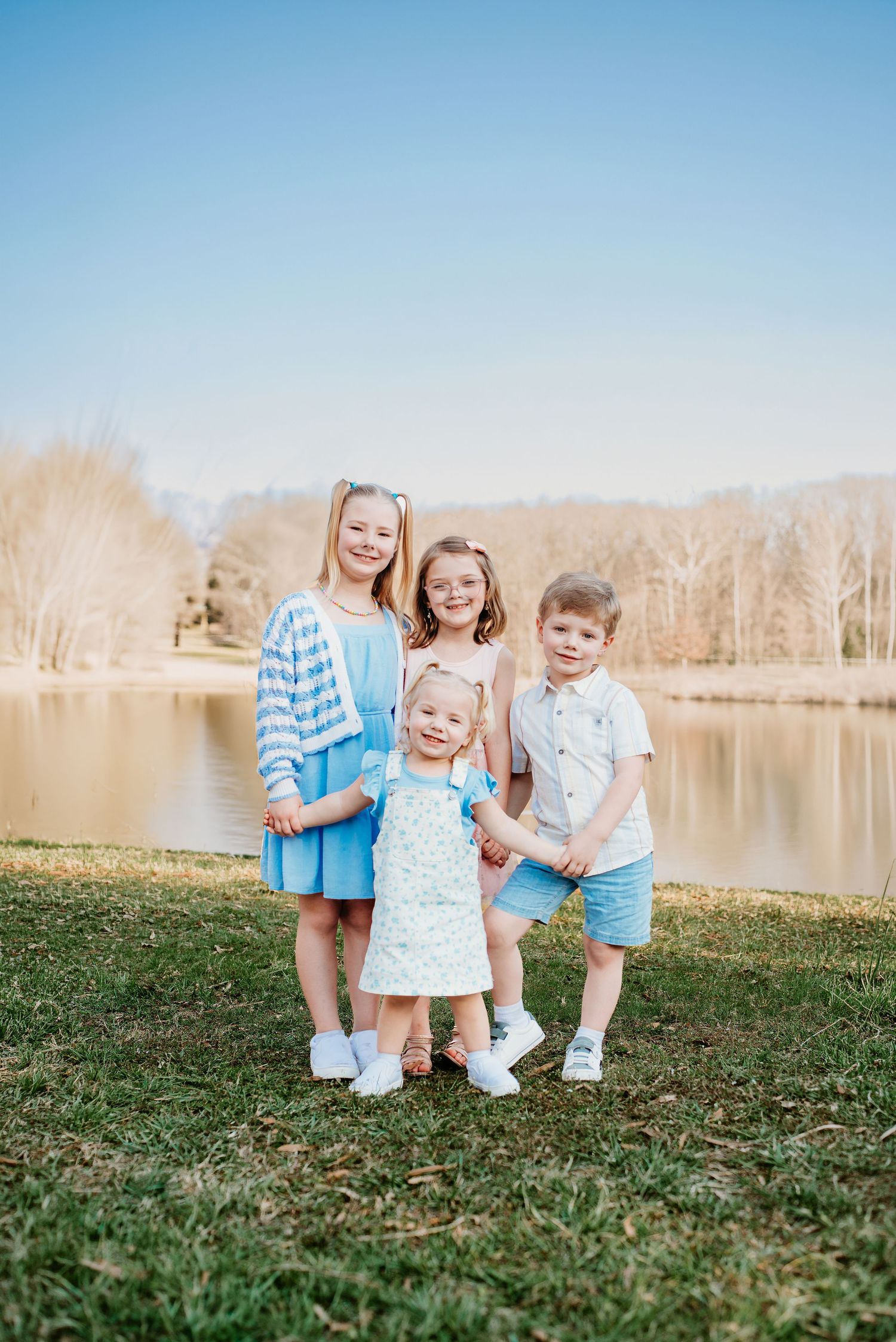 Four young children smiling together outdoors near a pond on a sunny day, dressed in spring pastel outfits.