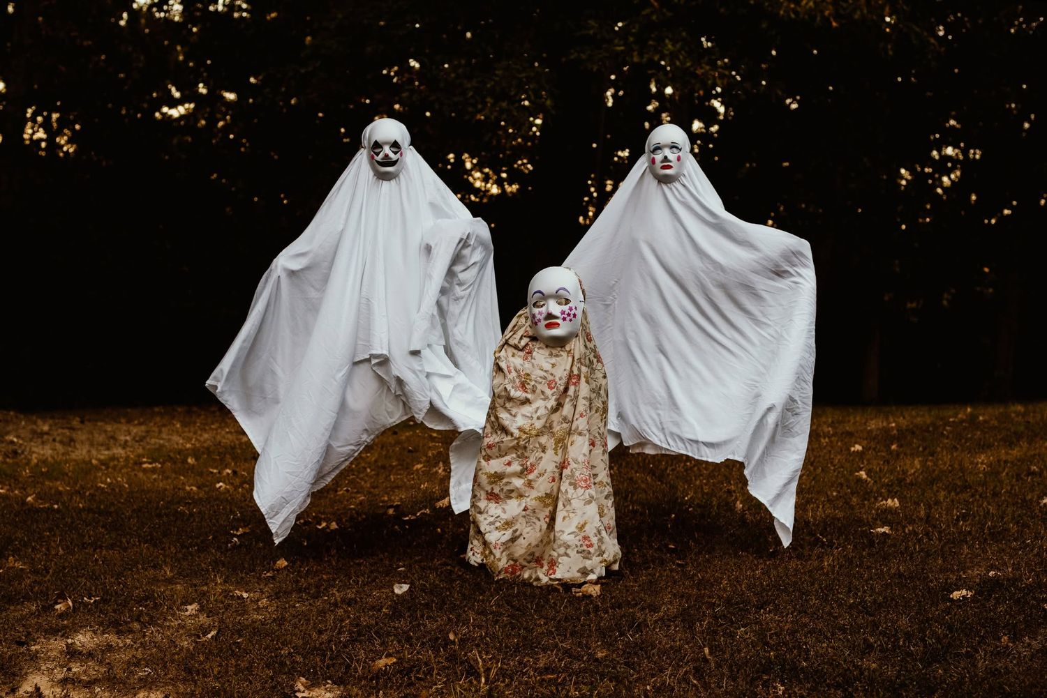 Three children in Halloween ghost costumes with white sheets and spooky masks standing outdoors near dark trees.