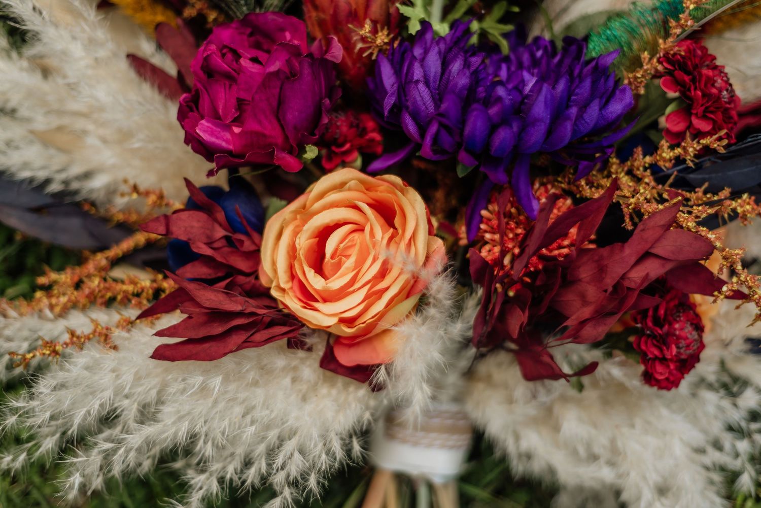Vibrant wedding bouquet featuring orange roses, purple flowers, burgundy dahlias, and fluffy pampas grass accents.