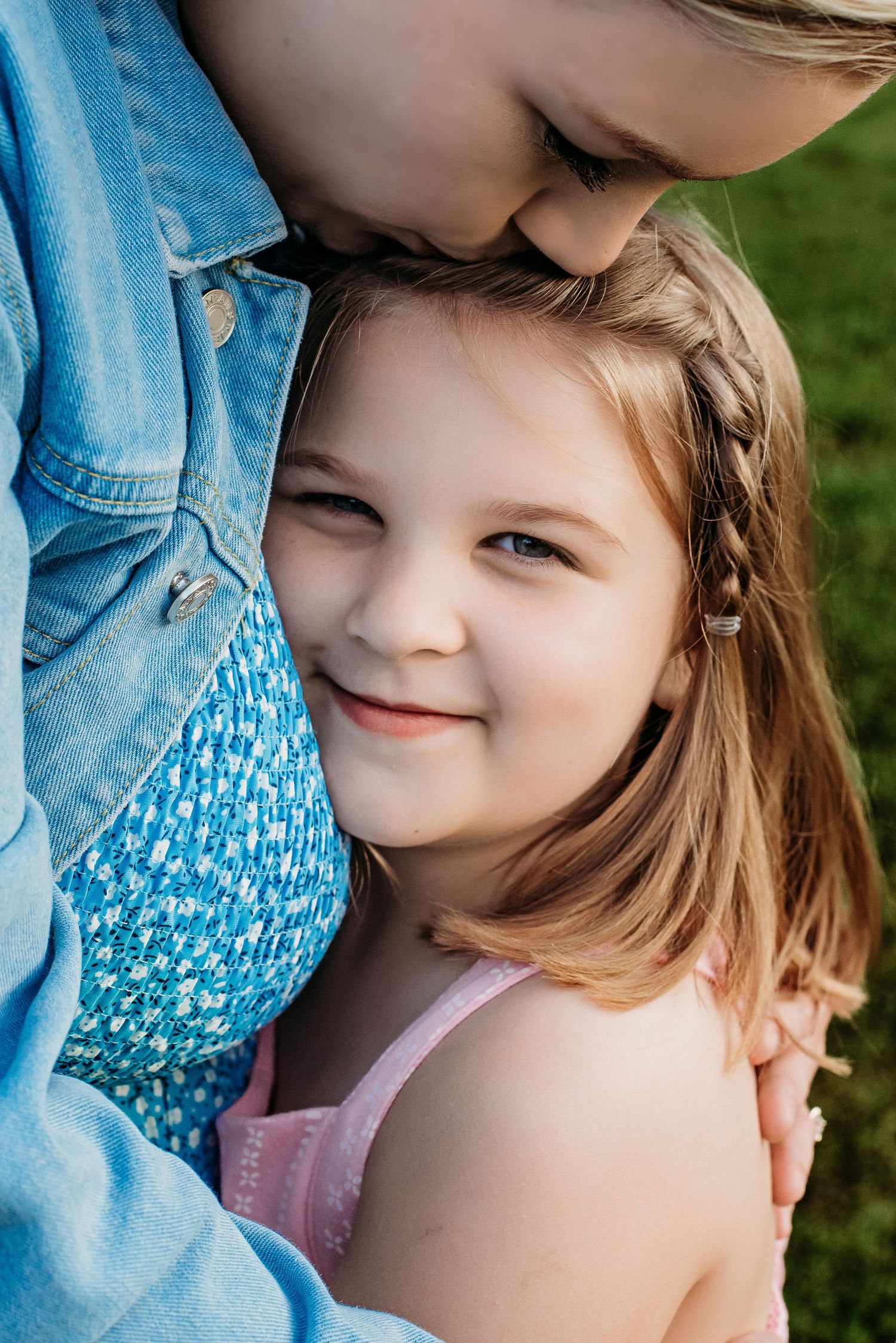 A young girl smiles warmly while snuggled against an older child outdoors in a sweet sibling embrace.