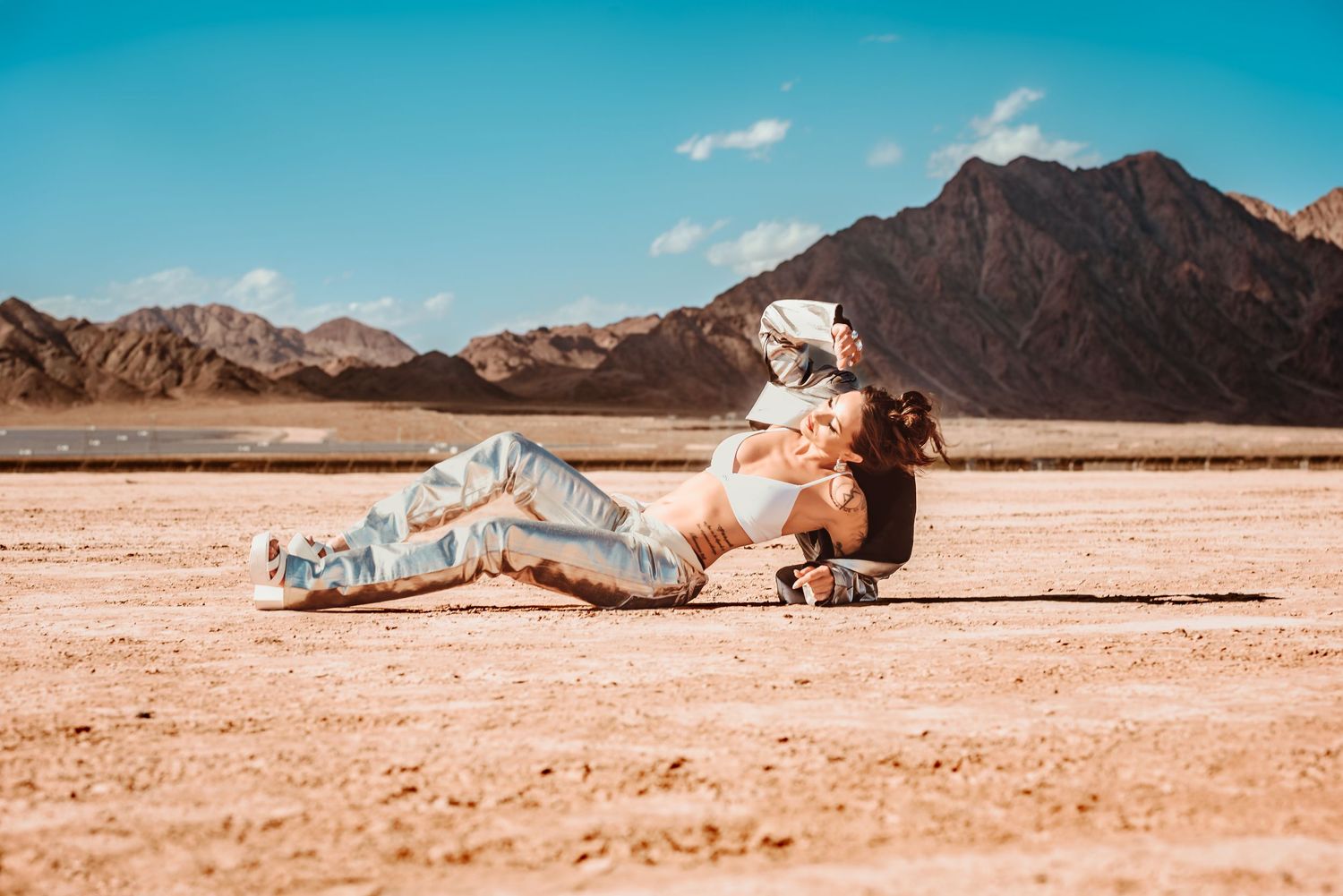 Person lying down in metallic outfit on desert sand with mountains in background.