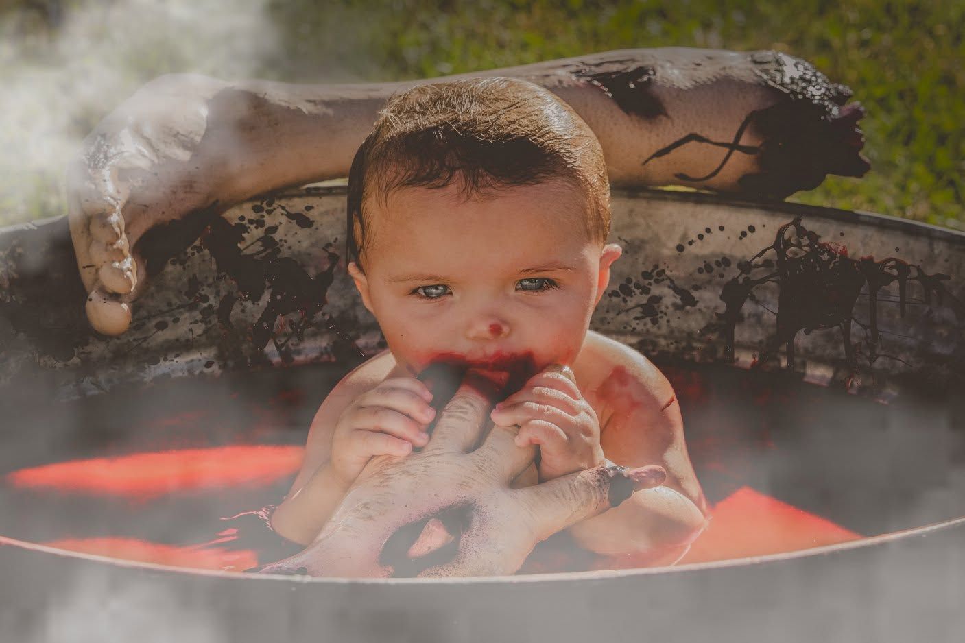 A baby boy sits in a steaming outdoor tub, chewing on fingers, surrounded by misty air and warm glowing light.