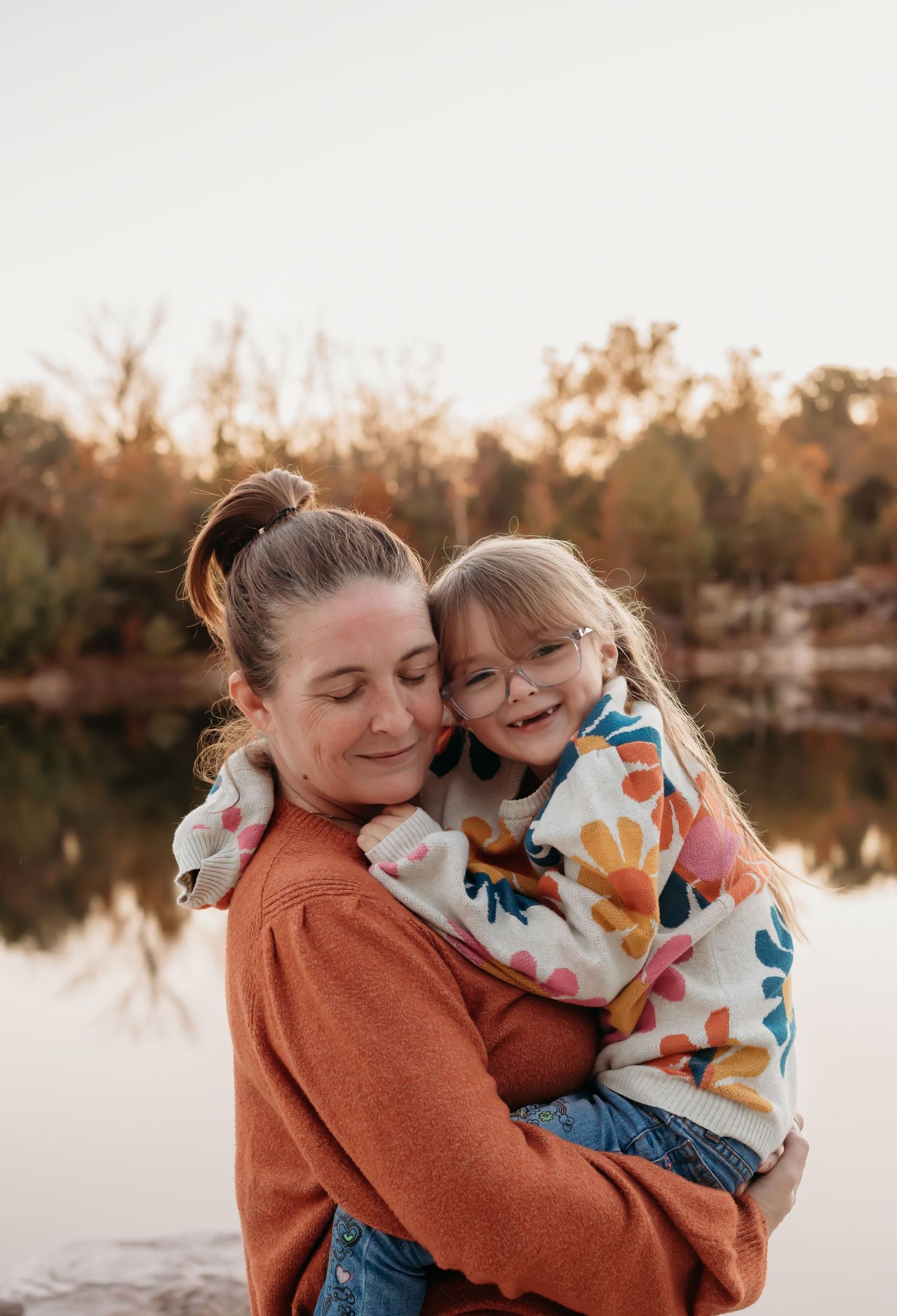Mother and child sharing a joyful embrace by a lake during golden hour sunset.