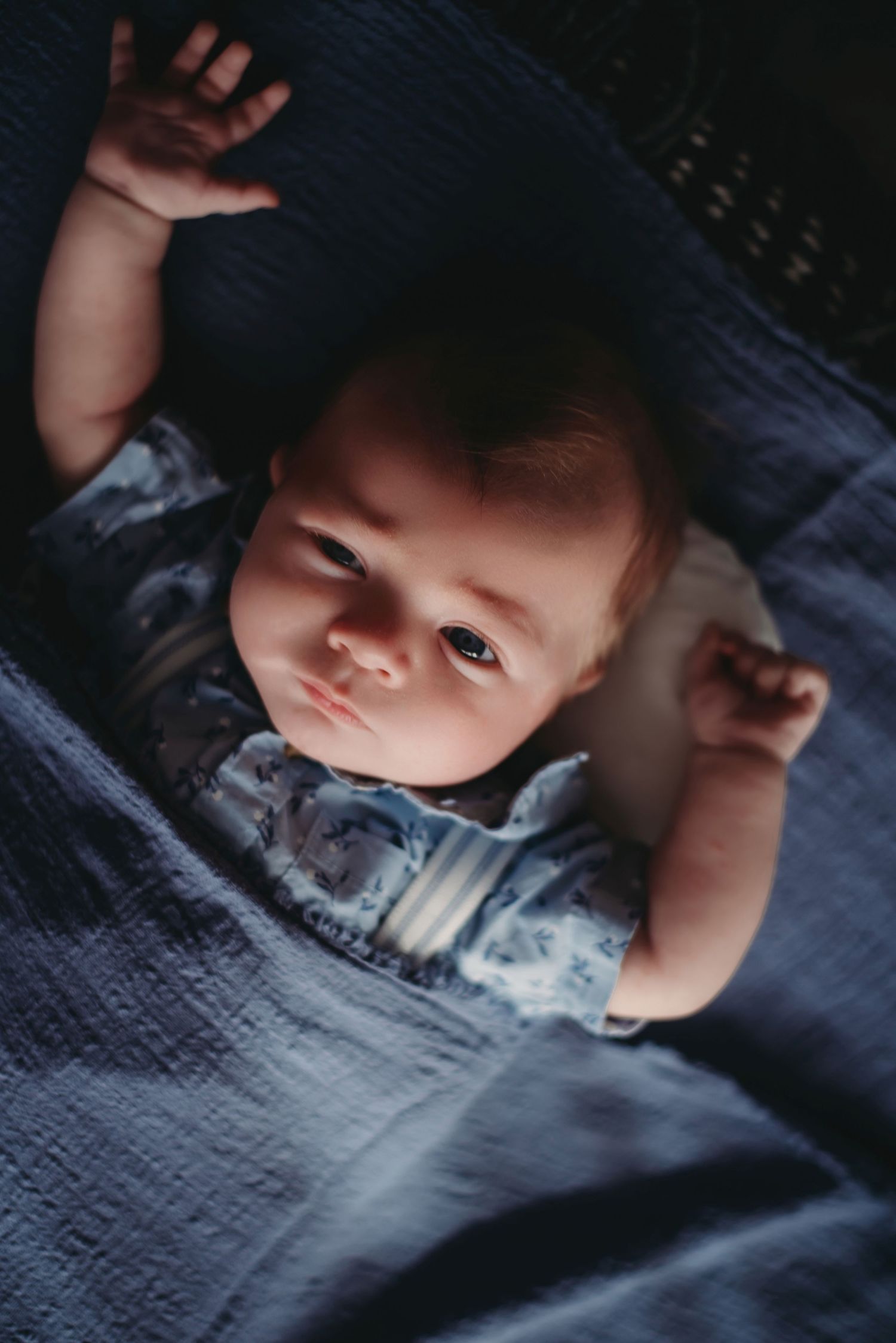 Baby lying on blue blanket looking up at camera in soft lighting.