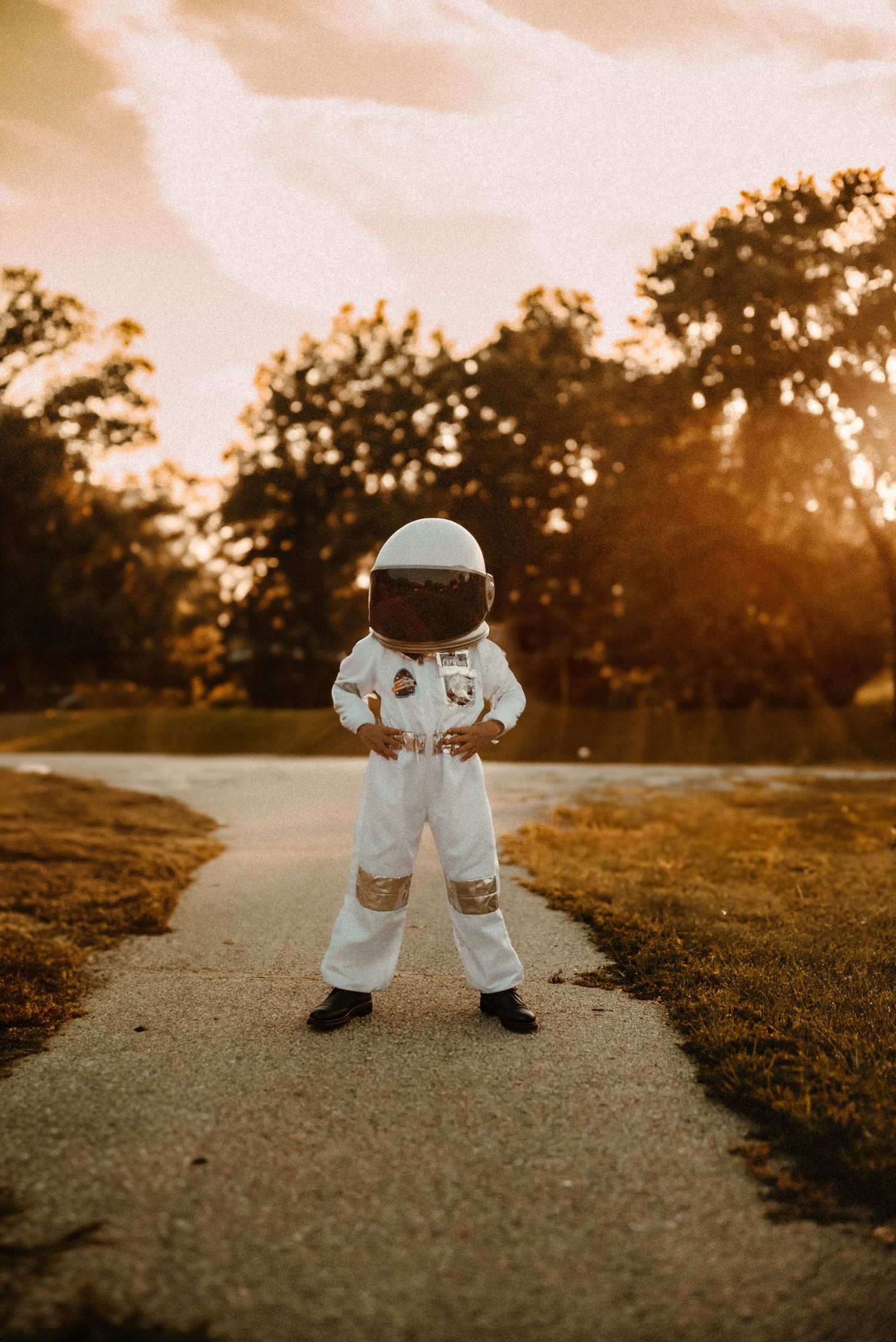 Small astronaut figure stands on path during golden sunset with trees silhouetted in background.
