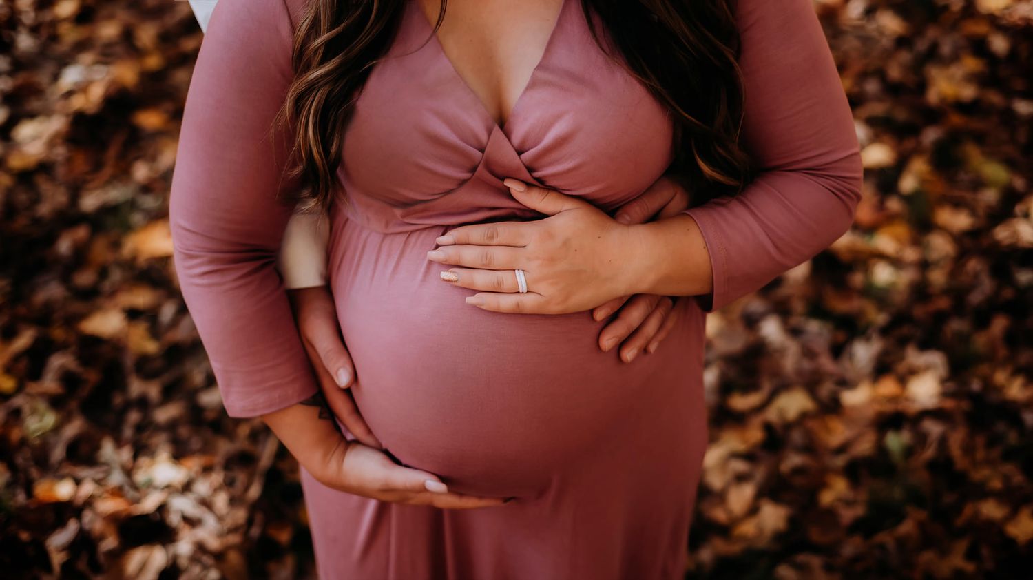 Close up of pregnant belly in mauve dress with hands cradling bump against fall leaves background.