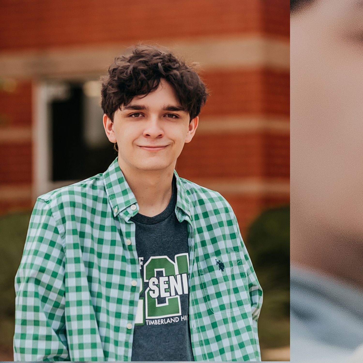 Student wearing green checkered shirt and gray t-shirt poses for casual portrait series against brick wall.