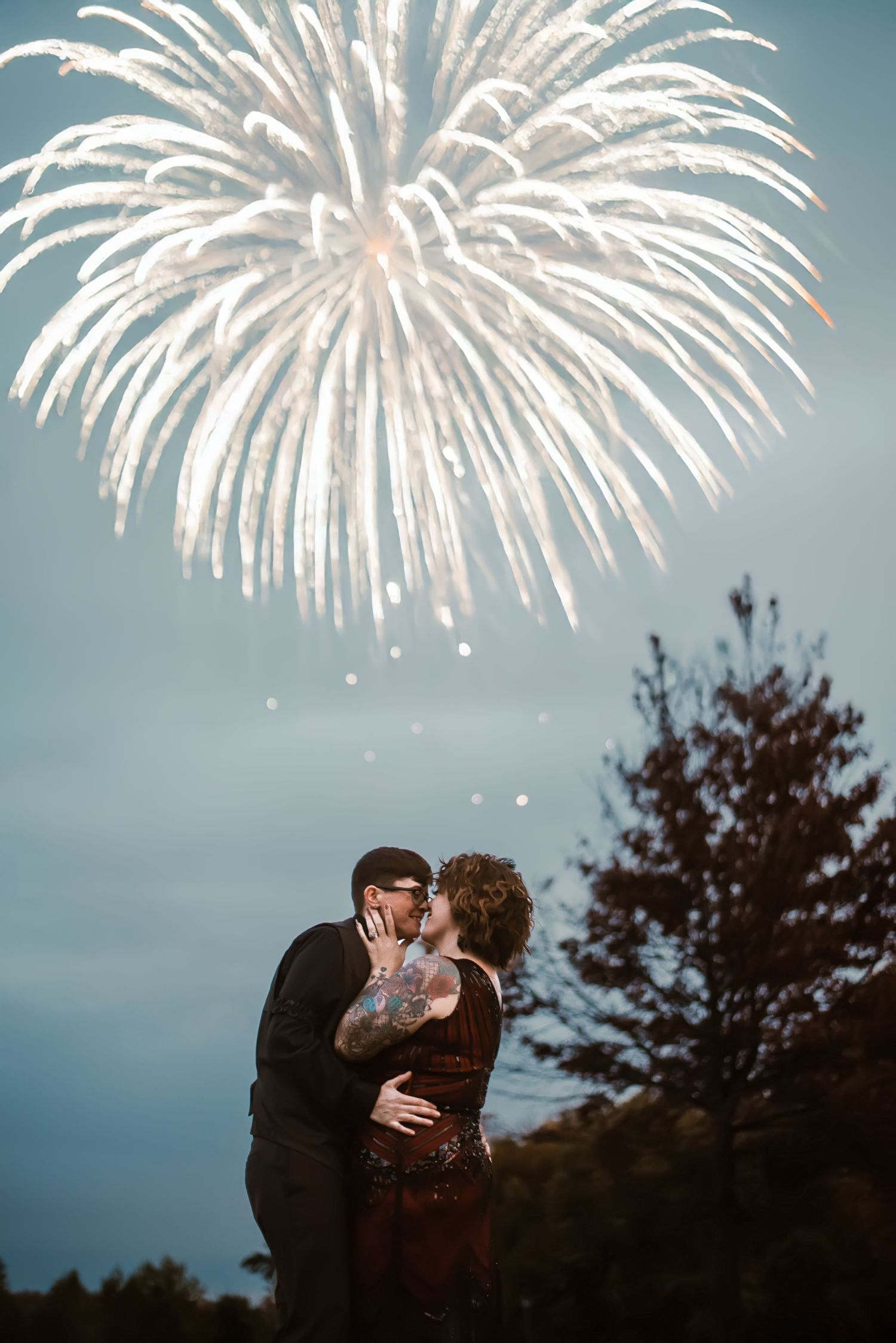 Couple kissing romantically under a stunning white fireworks display in the evening sky, surrounded by silhouetted trees.