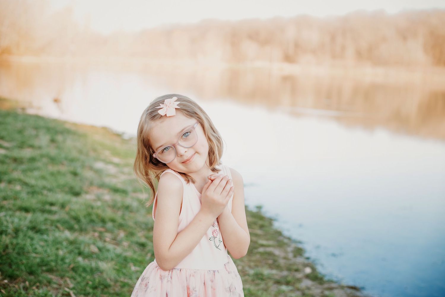 Young girl in pink dress and glasses smiling by a sunlit lake, wearing a floral hair clip.