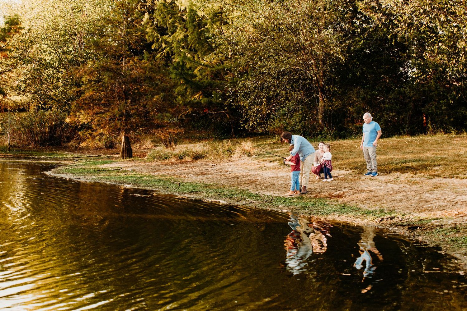 People enjoy recreational activities around a scenic pond on a sunny autumn day with golden foliage in the background.