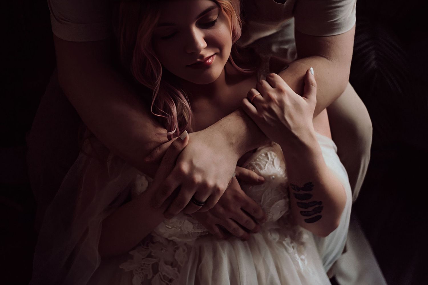 A bride in a lace wedding gown is tenderly embraced from behind in a moody, romantic portrait.