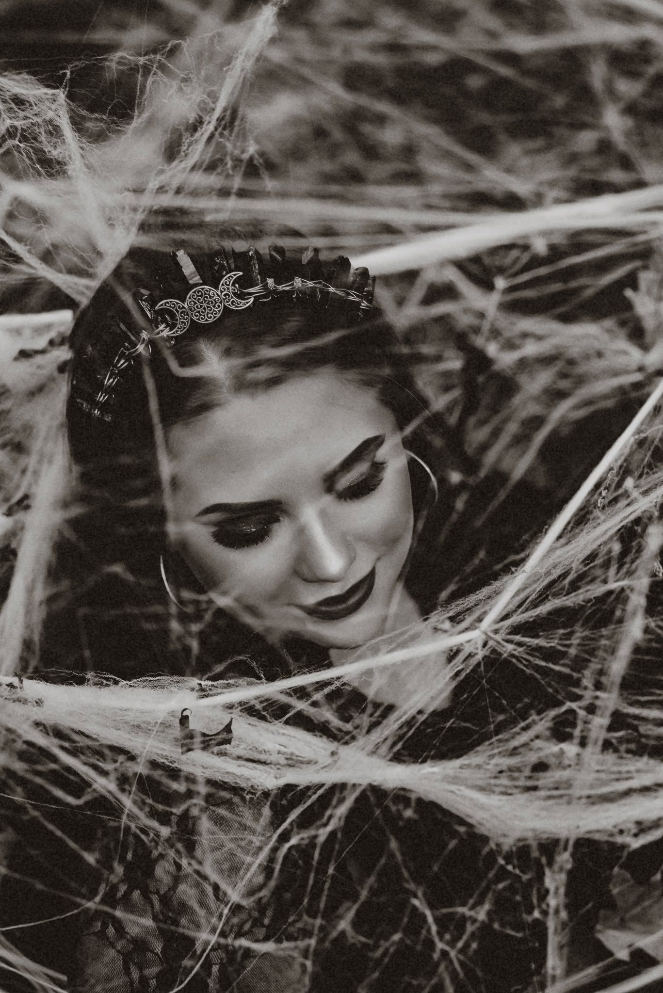 Black and white photo of a woman in a decorative crown surrounded by dramatic spider webs in a Halloween-themed portrait.