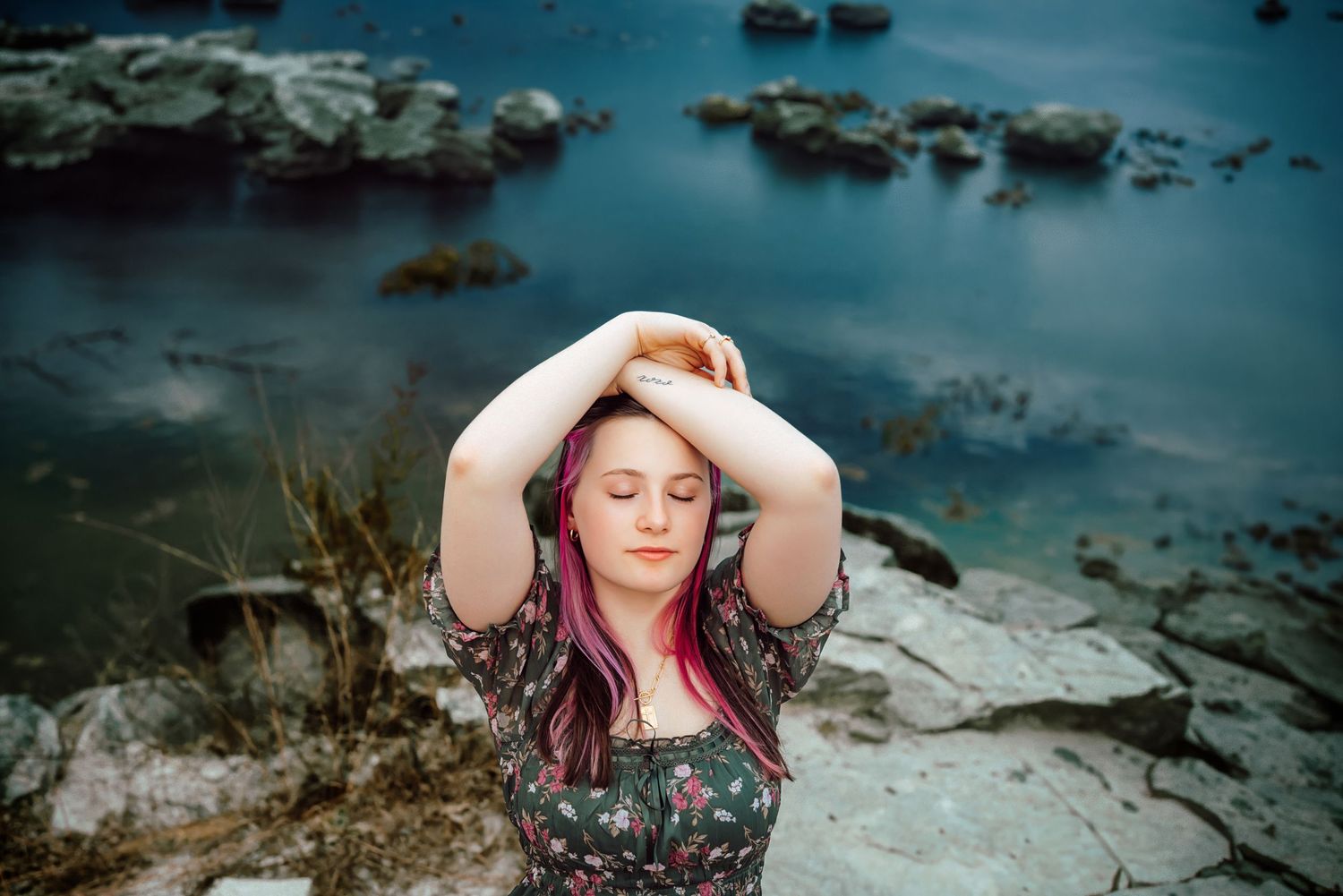 Young woman with pink hair poses by a rocky shoreline, hands behind her head, eyes closed peacefully.