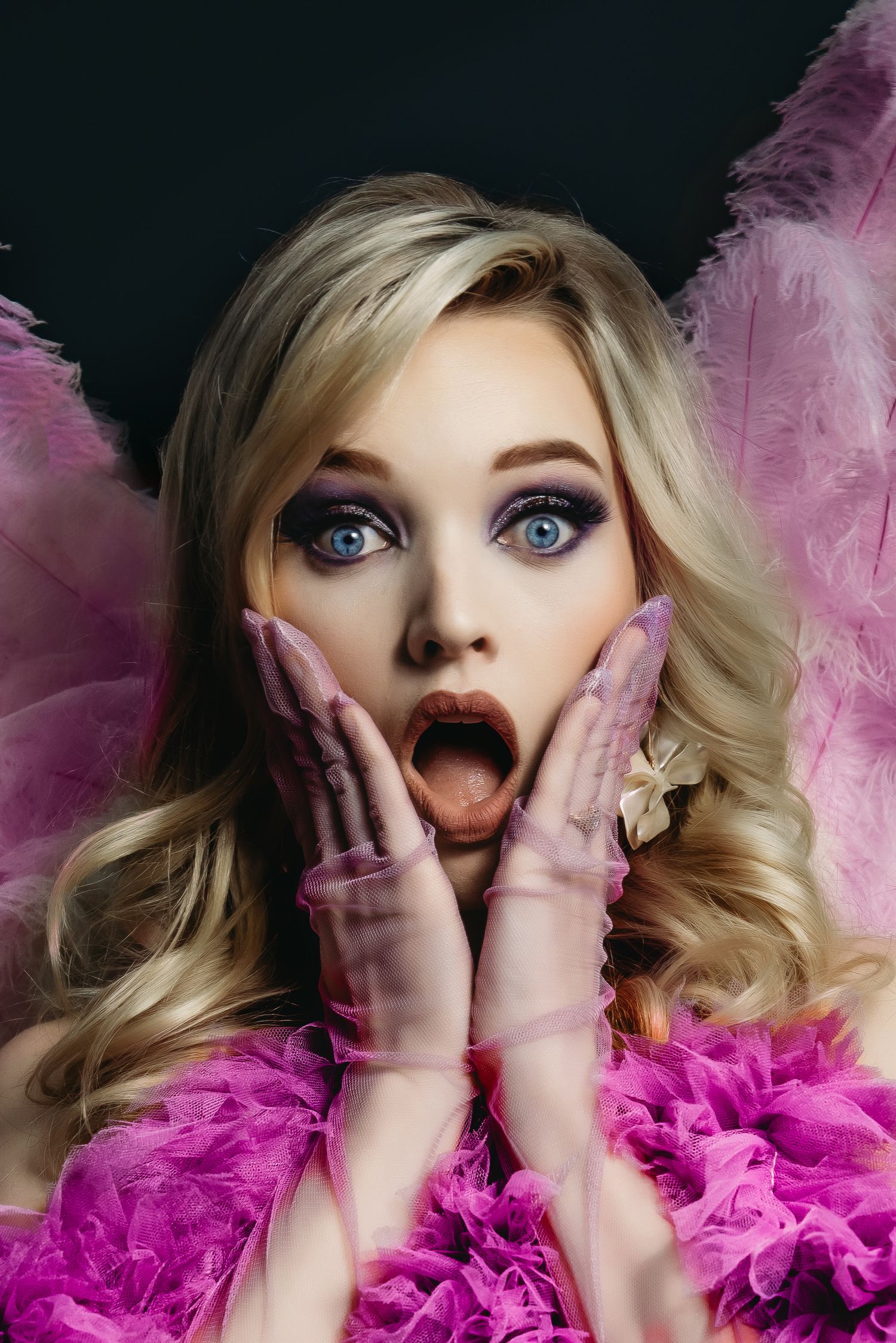 Dramatic portrait with pink feather boa and surprised expression against dark background.