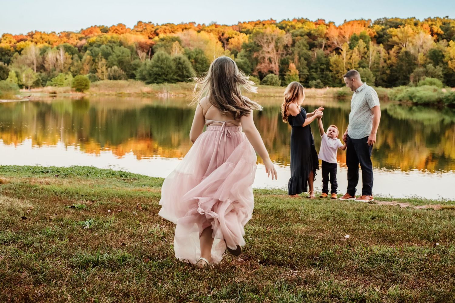 Girl in pink dress runs toward family by autumn lake with colorful fall foliage reflected in water.