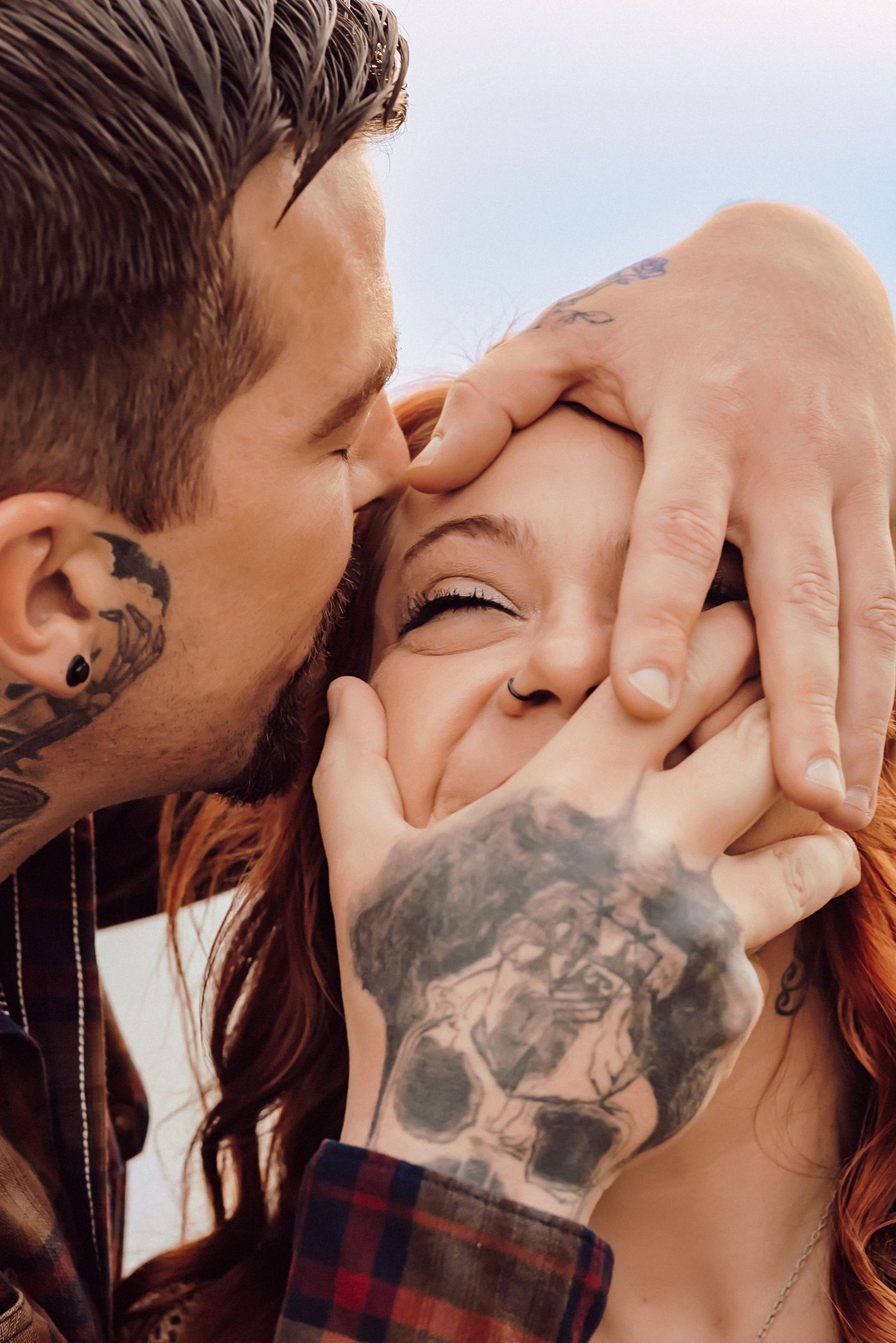 A tattooed couple sharing a tender kiss, with the man gently cupping the laughing woman's face with his tattooed hand.