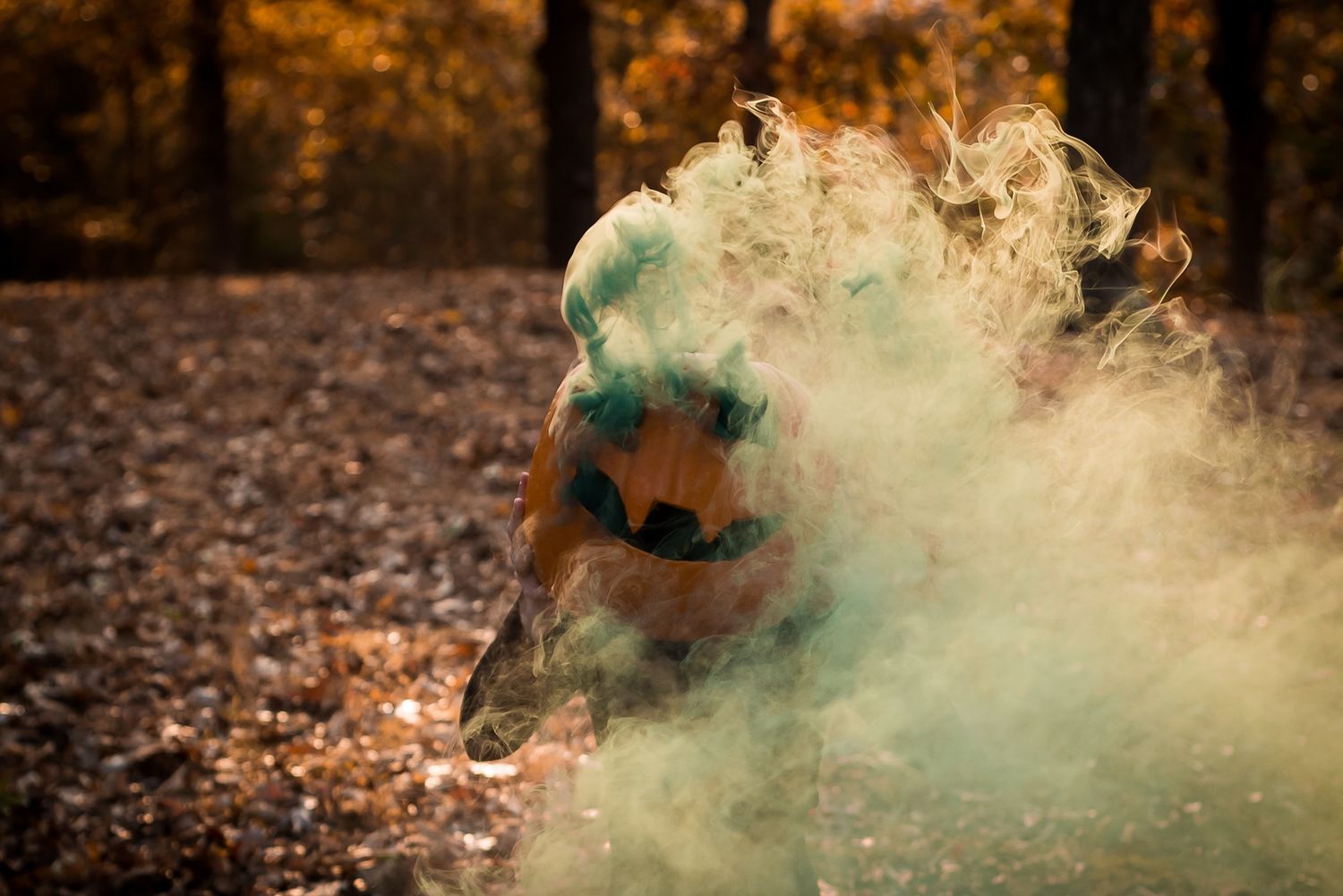 Green smoke billows around a carved pumpkin in a moody autumn forest scene.