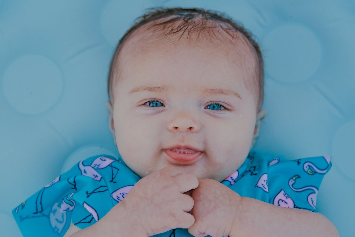 A baby wearing blue rests on light blue bedding.