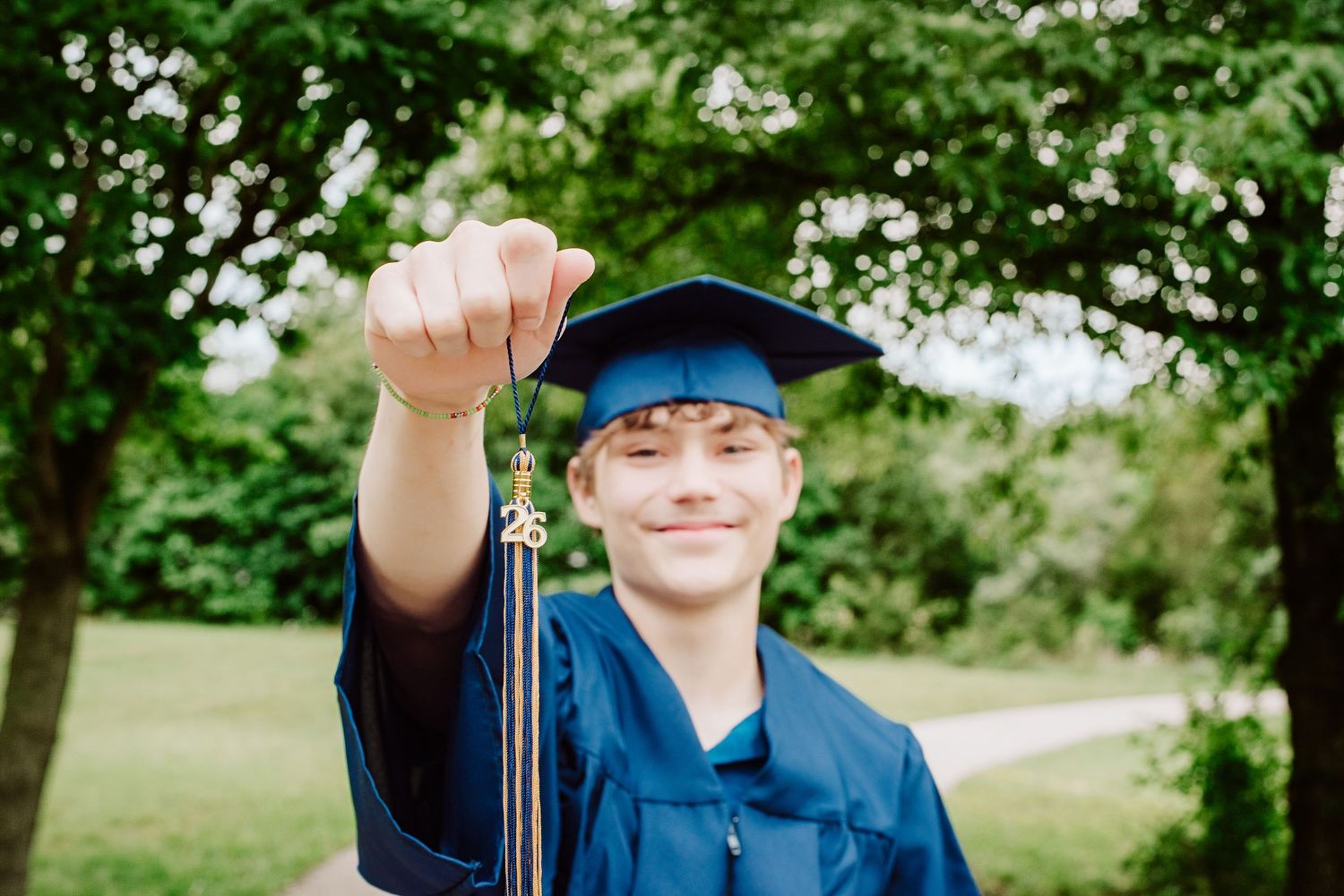 Smiling graduate in blue cap and gown holding tassel outdoors among green trees.